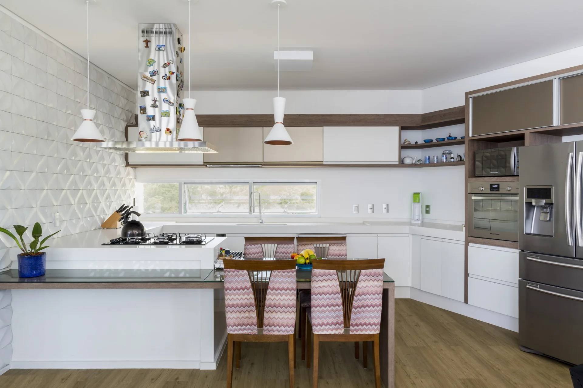 A modern white kitchen features a wood-accented island with an integrated glass-topped dining table and four chevron-patterned chairs.