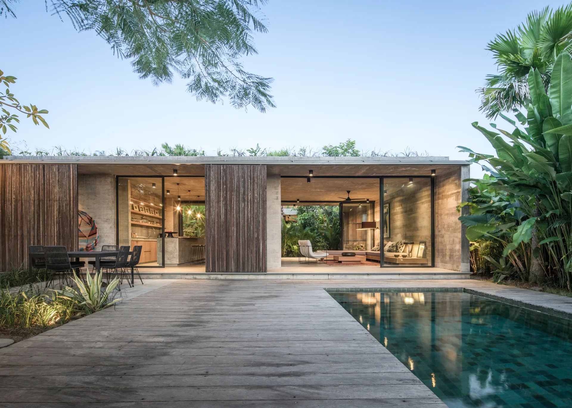 A tropical modern concrete residence with an outdoor swimming pool, wooden deck, and warm illuminated interiors visible through sliding glass doors.