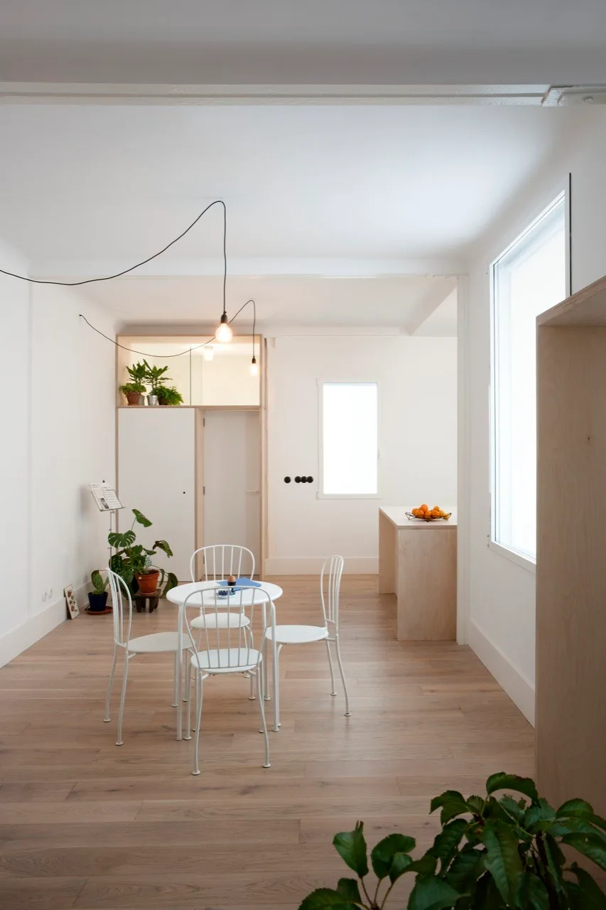 A minimalist white dining room features a round table with four metal chairs on light wood flooring near a built-in partition.
