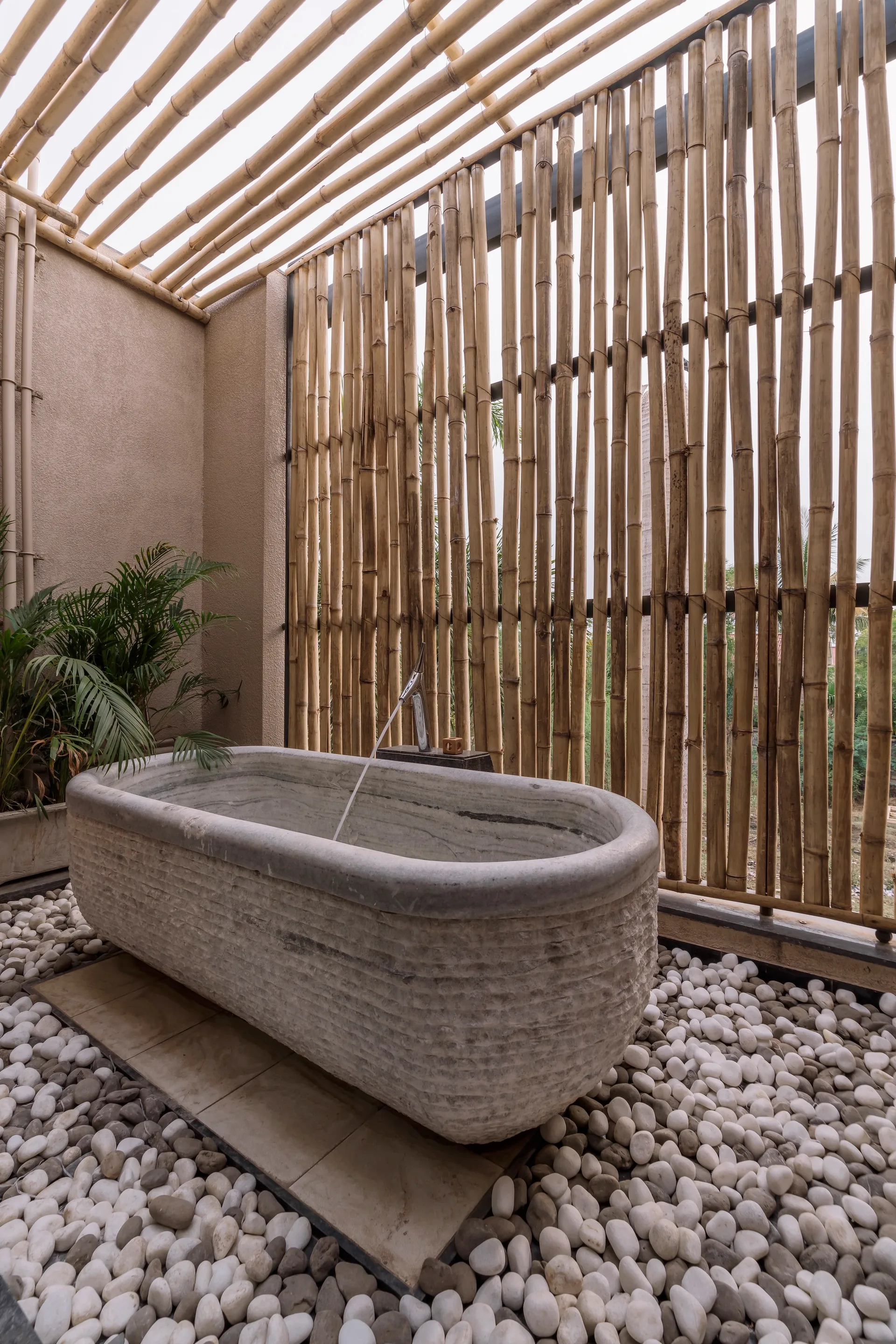 A carved stone bathtub with running water sits on a floor of white pebbles surrounded by vertical bamboo privacy screens and a bamboo slat ceiling.