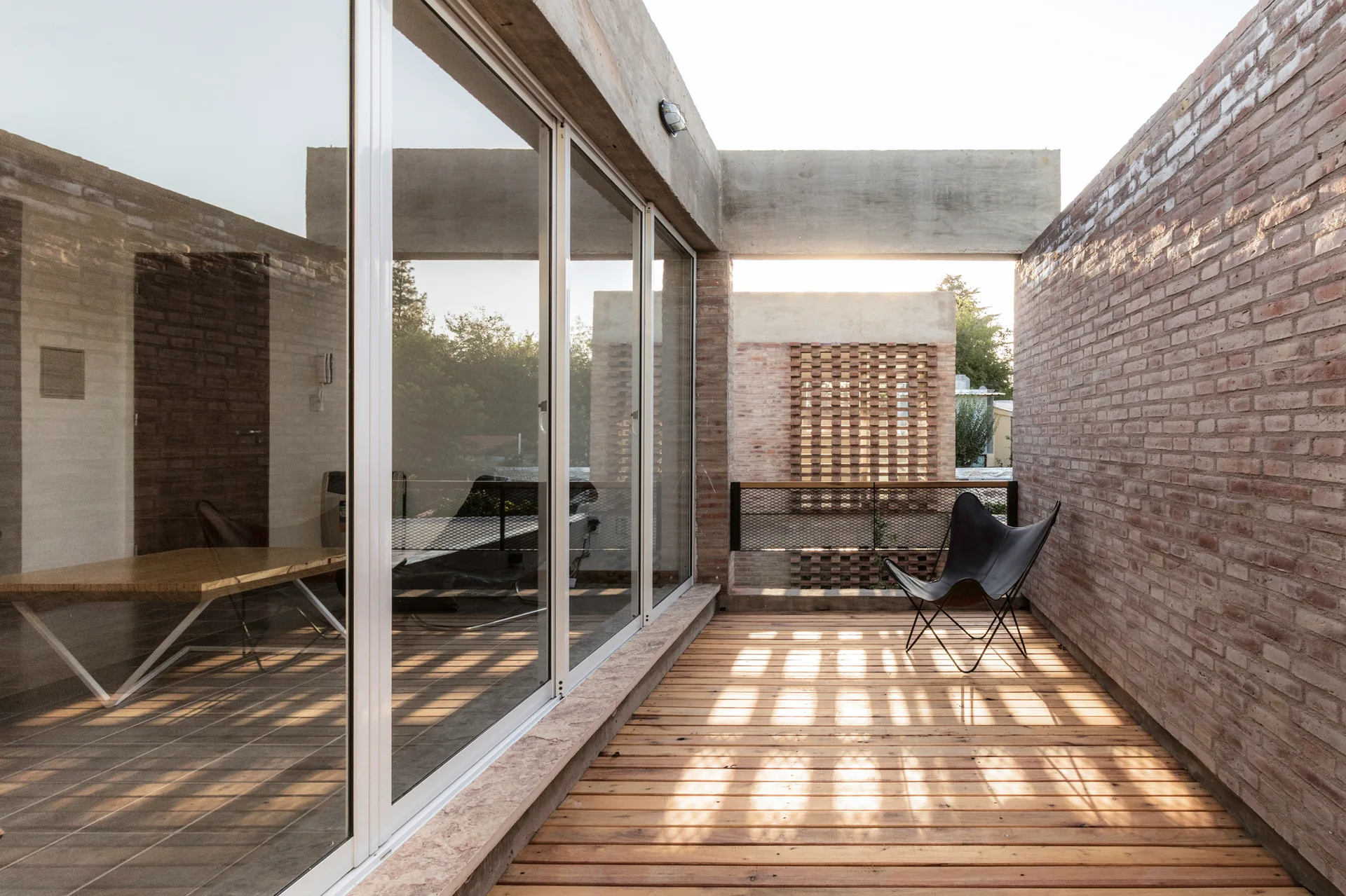 An outdoor balcony features warm wood deck flooring, textured brick walls, a black metal mesh railing, and a classic black butterfly chair.