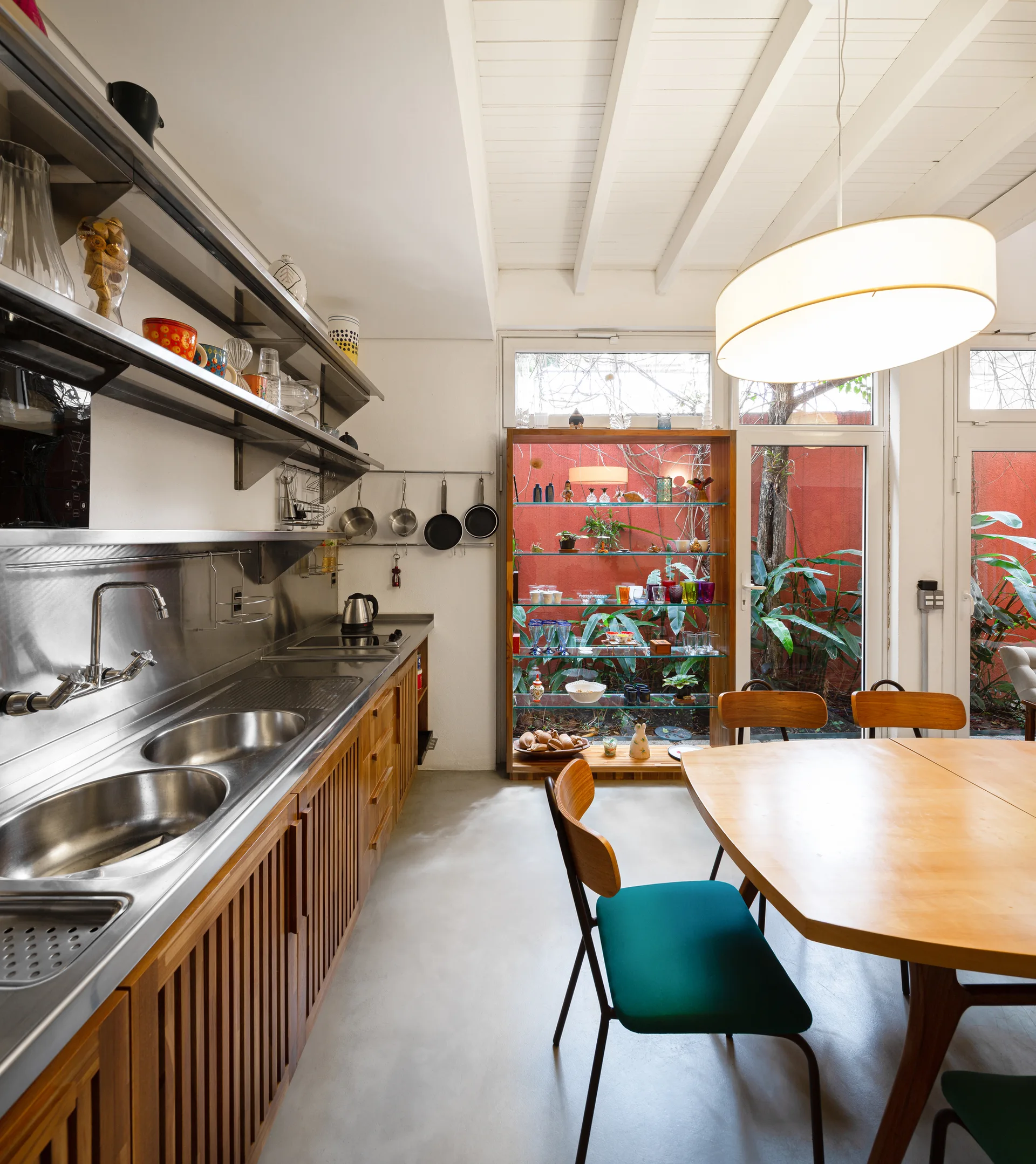 A kitchen and dining area with slatted wood cabinetry, a stainless steel sink, and glass doors looking out onto a lush courtyard garden.