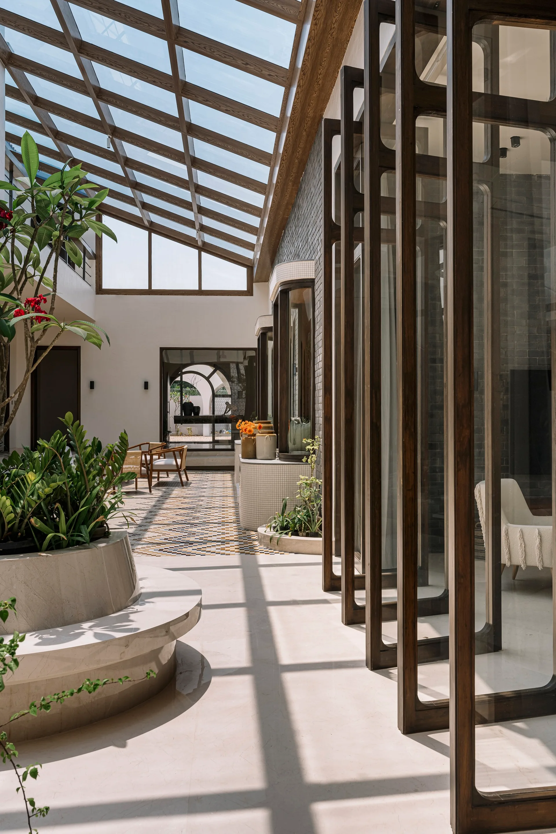 An indoor courtyard features a pitched glass skylight, tall dark wood framed glass doors, and a central stone planter bench on a polished stone floor.
