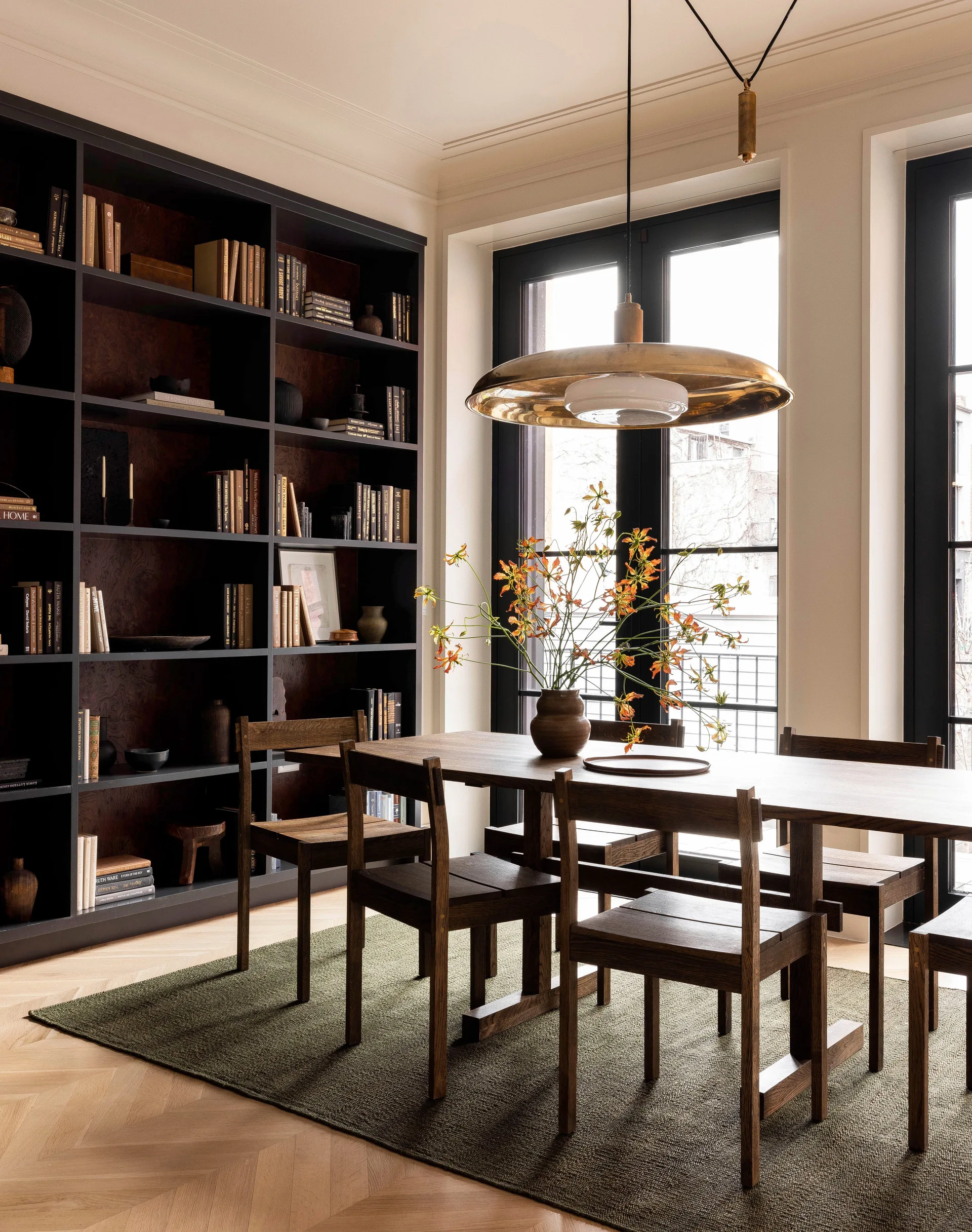 A dining room features a dark wood table and chairs on an olive green rug next to a black floor-to-ceiling bookshelf and tall windows.