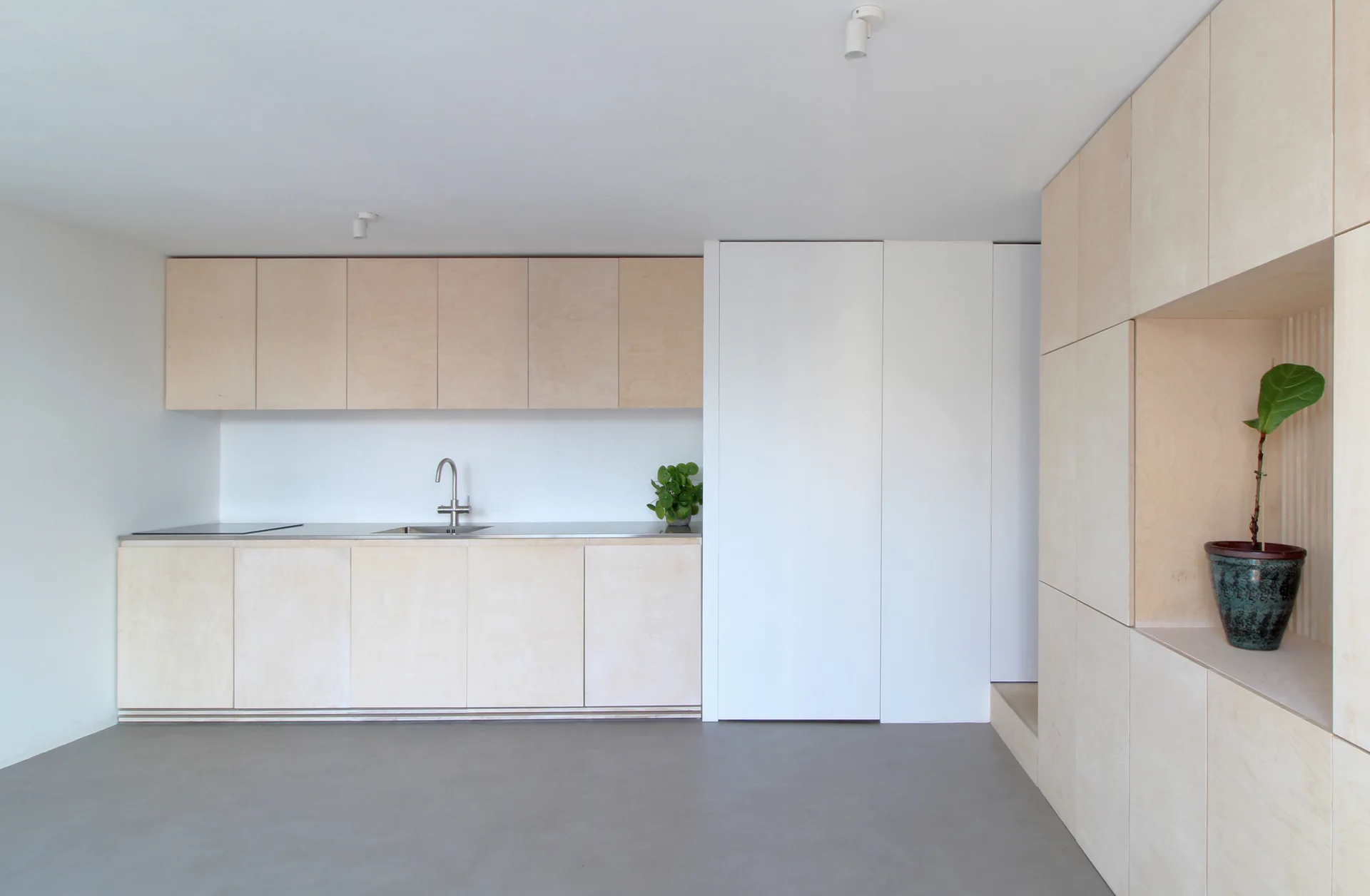 A minimalist kitchen features light plywood cabinetry, a stainless steel sink, a smooth gray floor, and a tall storage unit with a built-in niche.