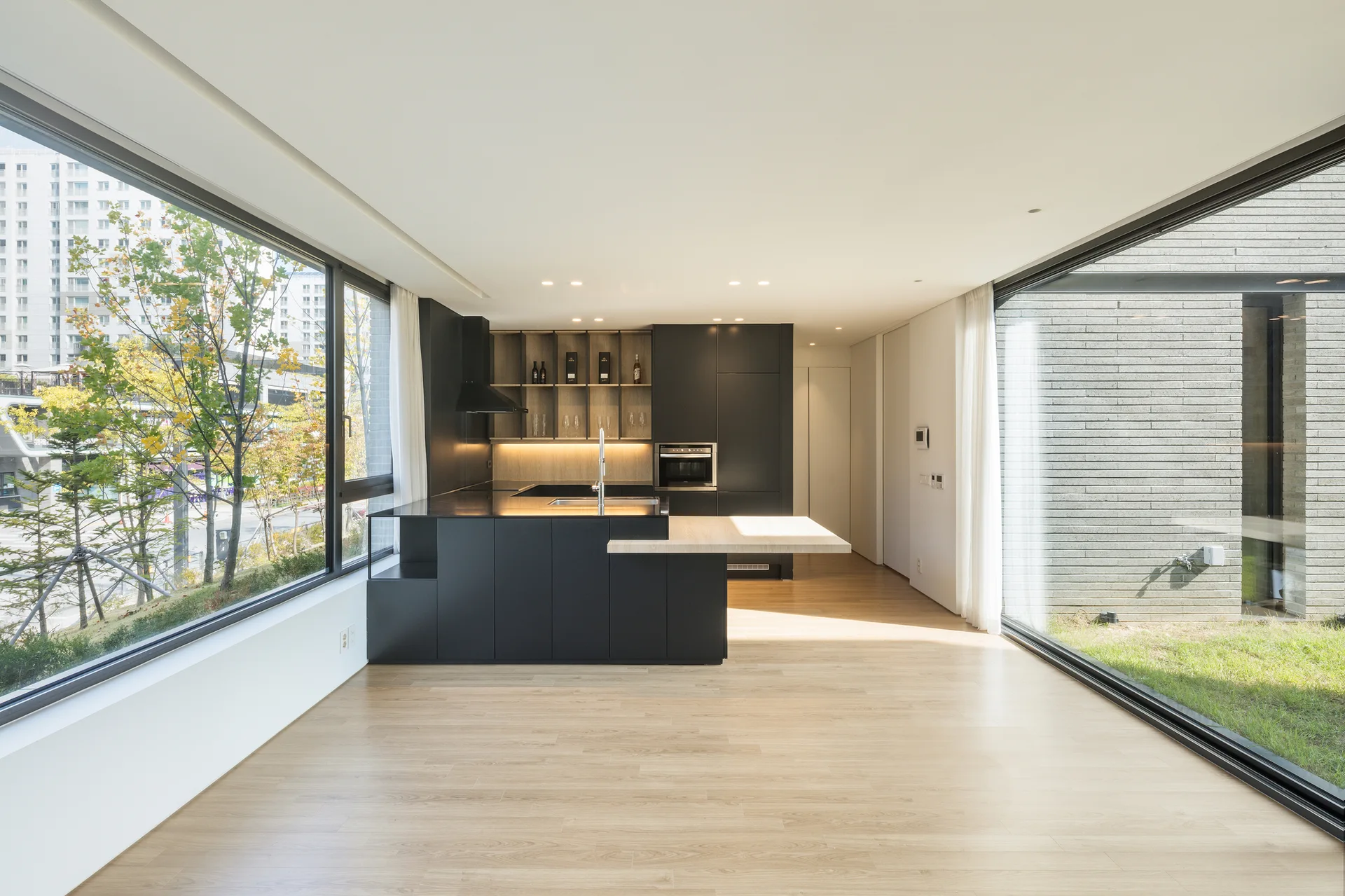 A minimalist kitchen features matte black cabinetry, light wood floors, and a kitchen island with an extended wood counter between large windows.
