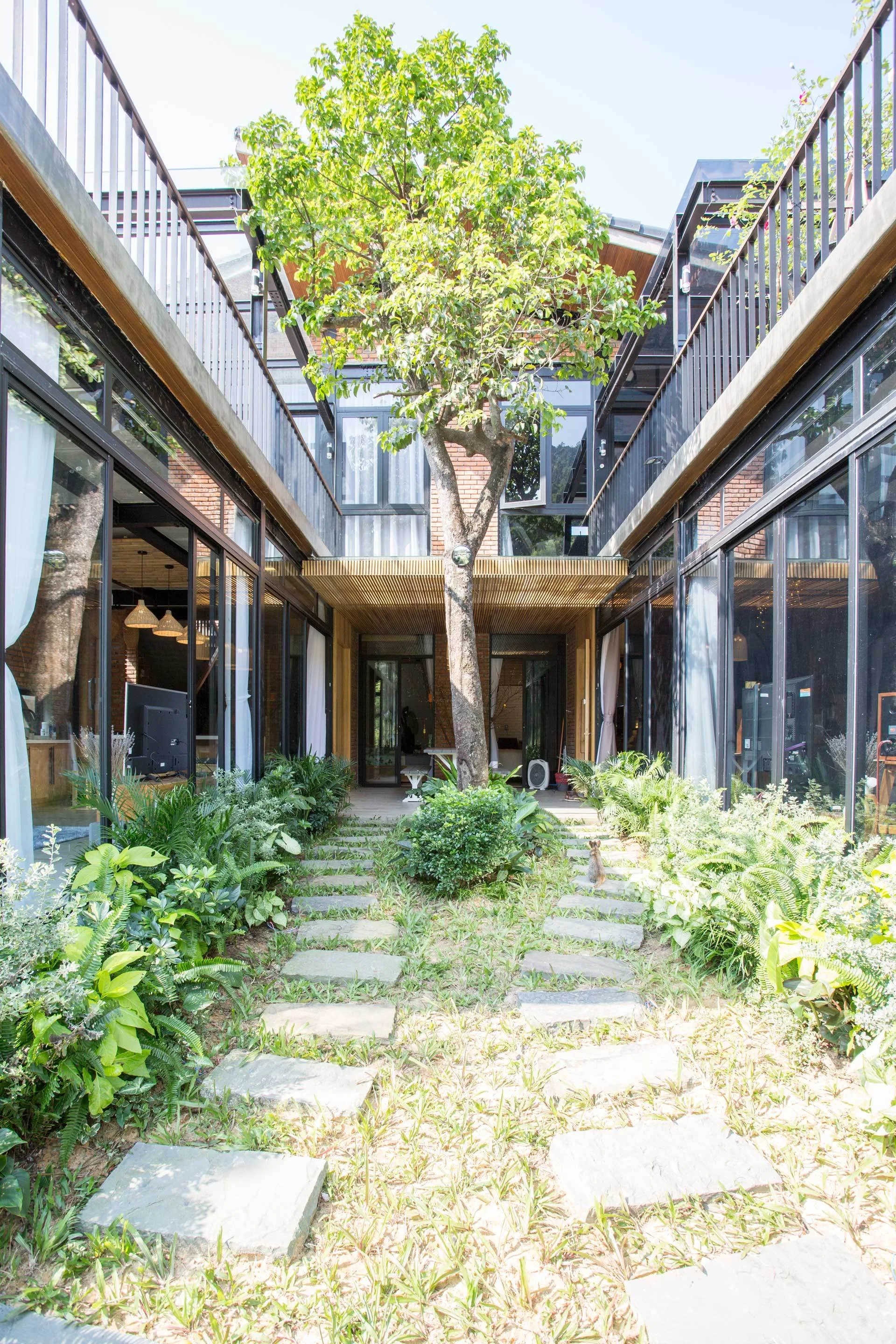A modern courtyard features a stepped stone path leading toward a central tree, framed by glass-walled residential wings with brick details.