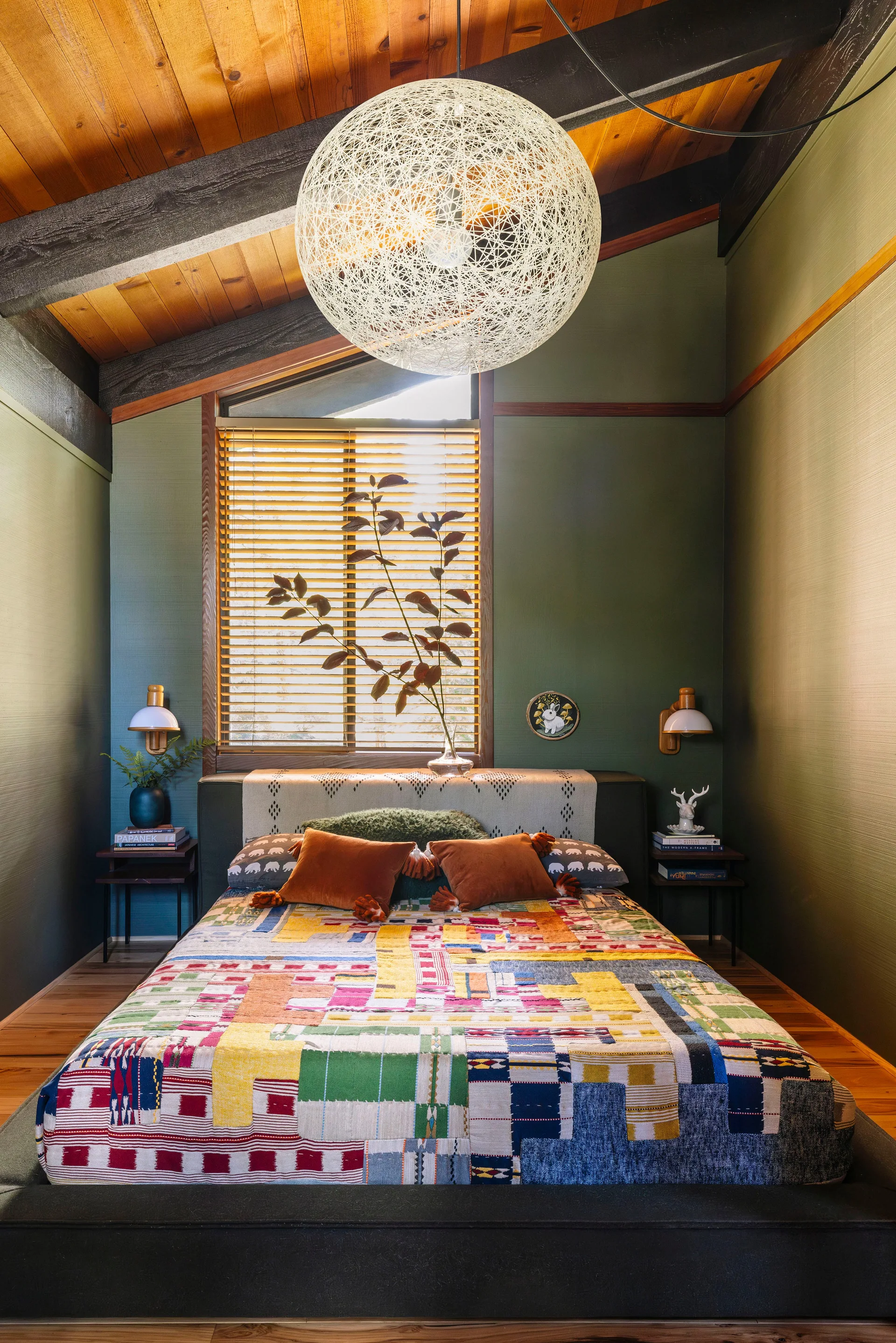 A mid-century bedroom features a colorful patchwork quilt on a low bed, muted green walls, and a vaulted wood plank ceiling with a large globe pendant.