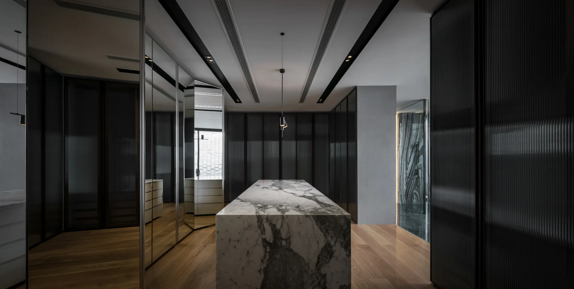 A minimalist dressing room features a central veined marble island, dark reeded glass storage cabinets, and mirrored closet doors on wood floors.