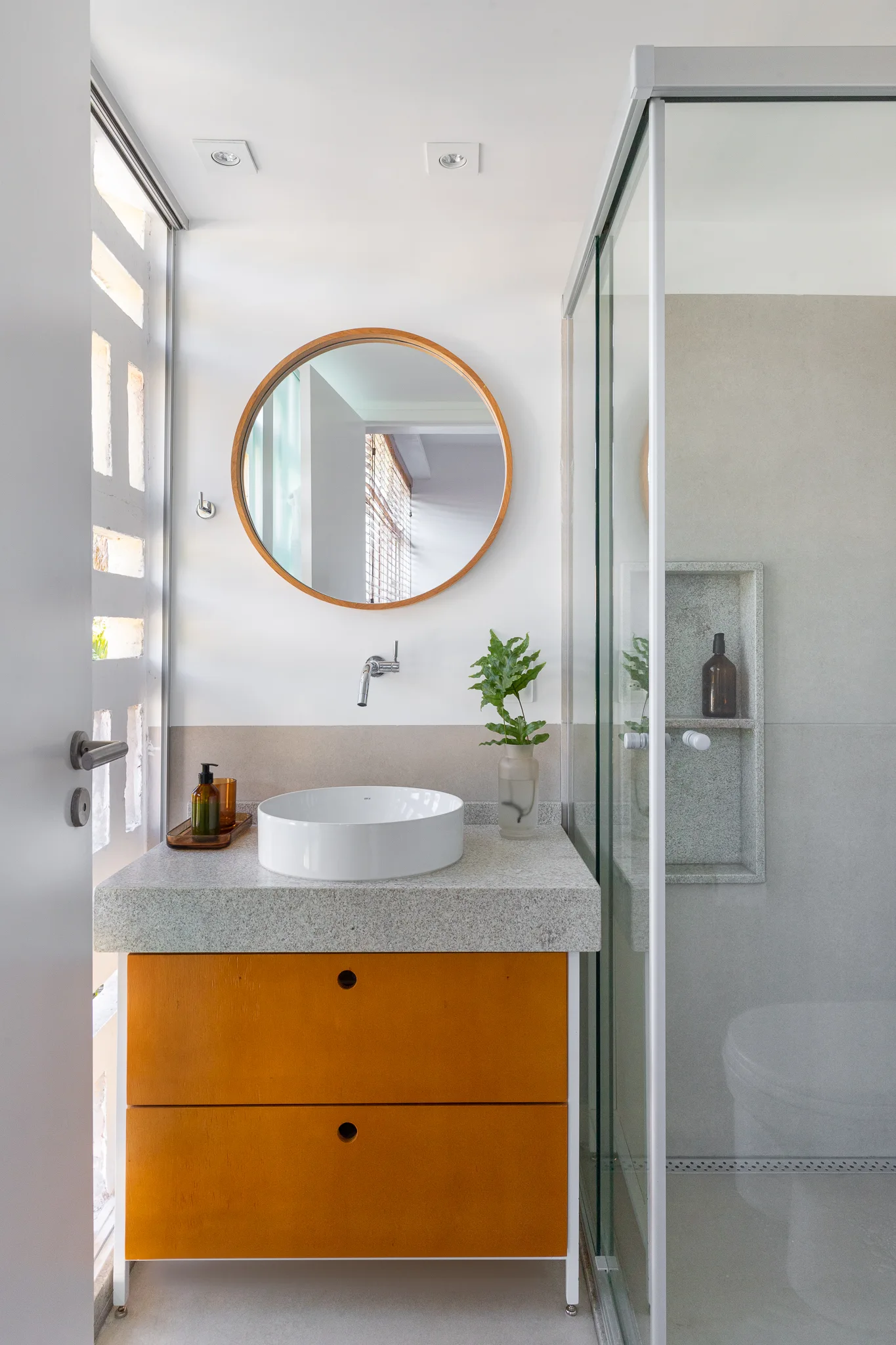 A modern bathroom features a round mirror over a white vessel sink on a speckled grey stone countertop with bright orange wooden drawers.