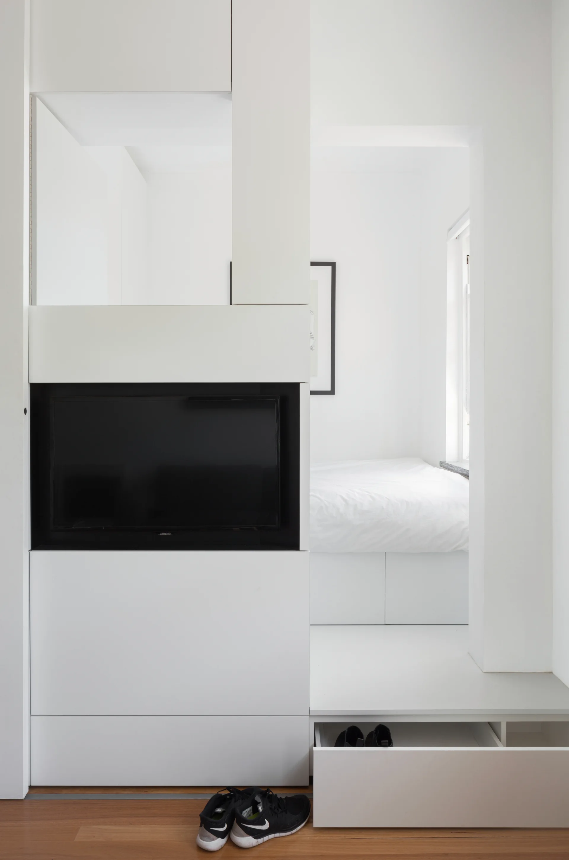 A minimalist bedroom features a white built-in storage unit containing a television next to an alcove with a platform bed and low pull-out drawer.