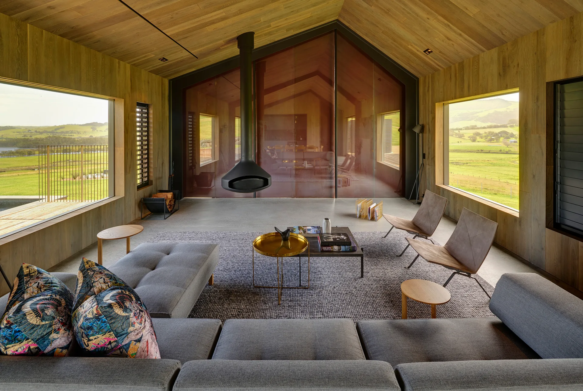 A modern living room features a black suspended fireplace, wood-clad walls and ceiling, a gray sectional, and windows overlooking hills.