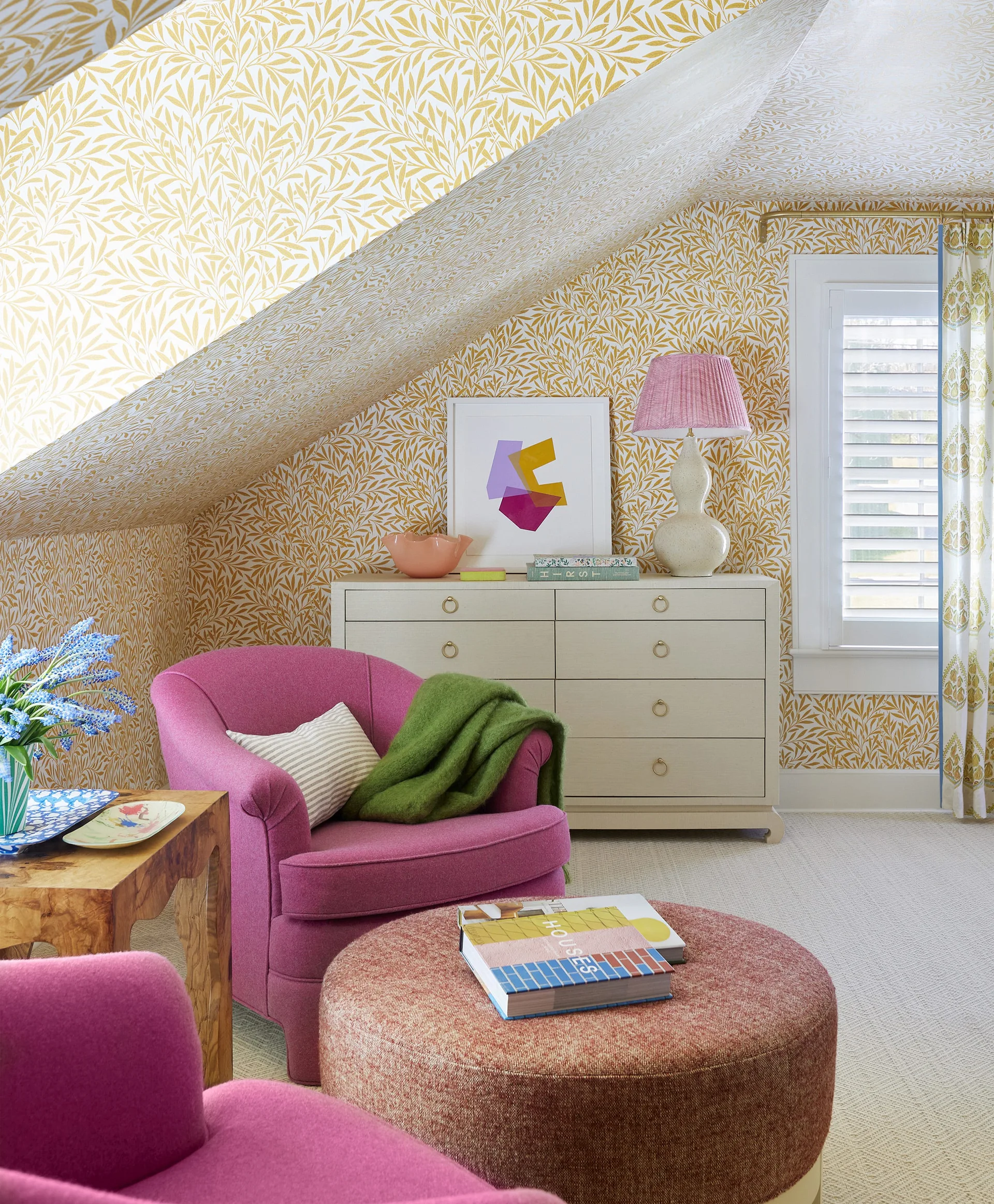 A bedroom corner under a sloped ceiling features yellow botanical wallpaper, two pink upholstered armchairs, and a cream dresser.