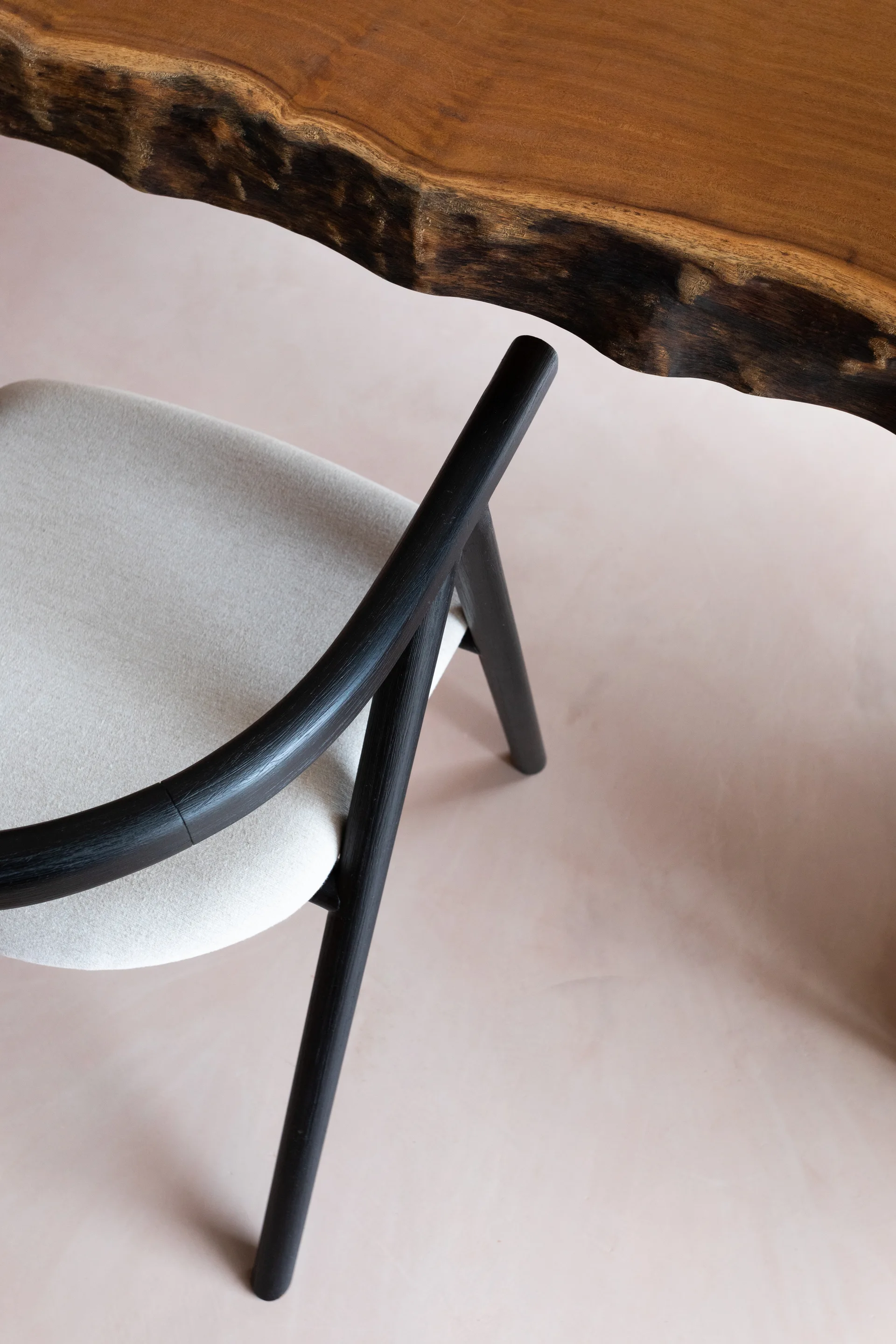 A black wooden dining chair with a light fabric seat sits beneath the curved border of a live-edge wood table on a smooth matte floor.
