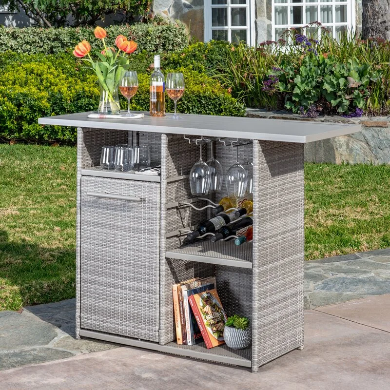 A grey woven wicker outdoor bar cart with a silver metal top features built-in wine storage on a stone patio.