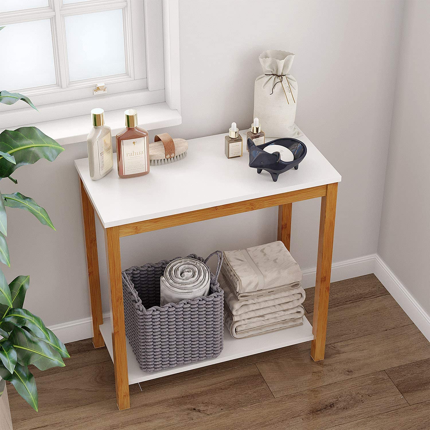 A two-tier white and wood console table sits in a bathroom corner under a window, holding toiletries, folded towels, and a grey knit basket.