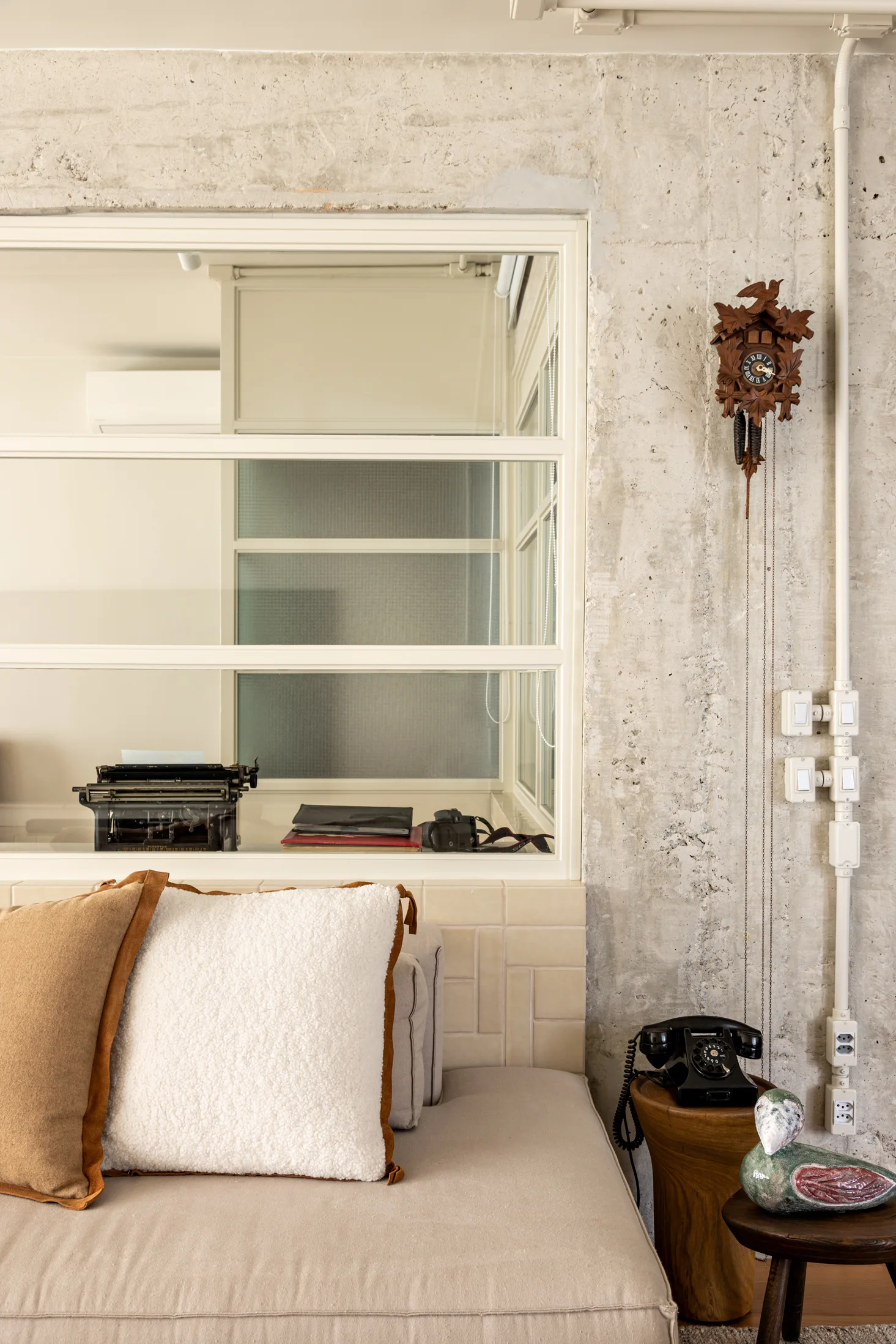 A beige sofa with textured cushions sits beneath an interior window and an exposed concrete wall decorated with a wooden cuckoo clock.