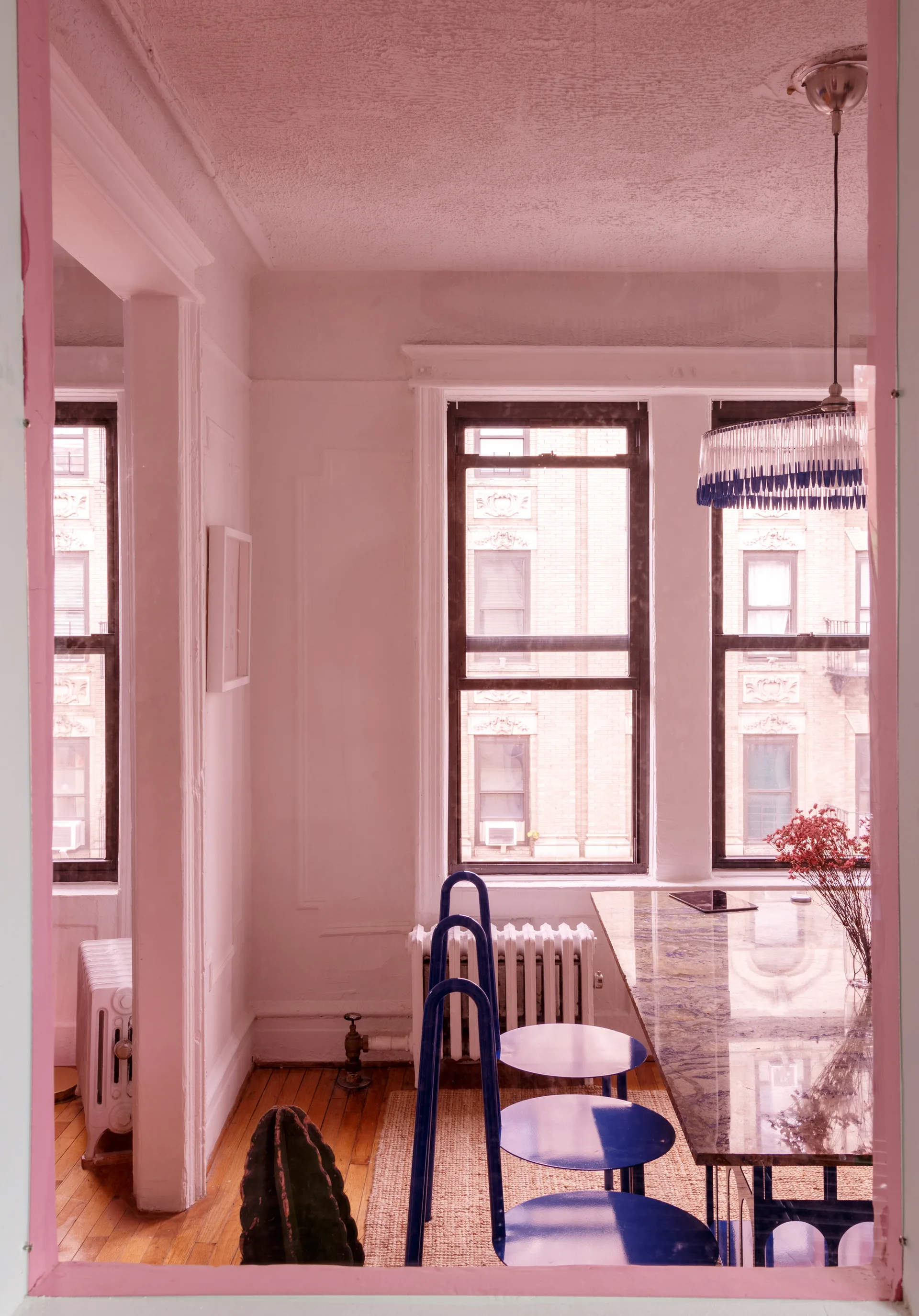 An apartment dining room features three cobalt blue chairs next to a long stone table under a fringed pendant light with urban windows.