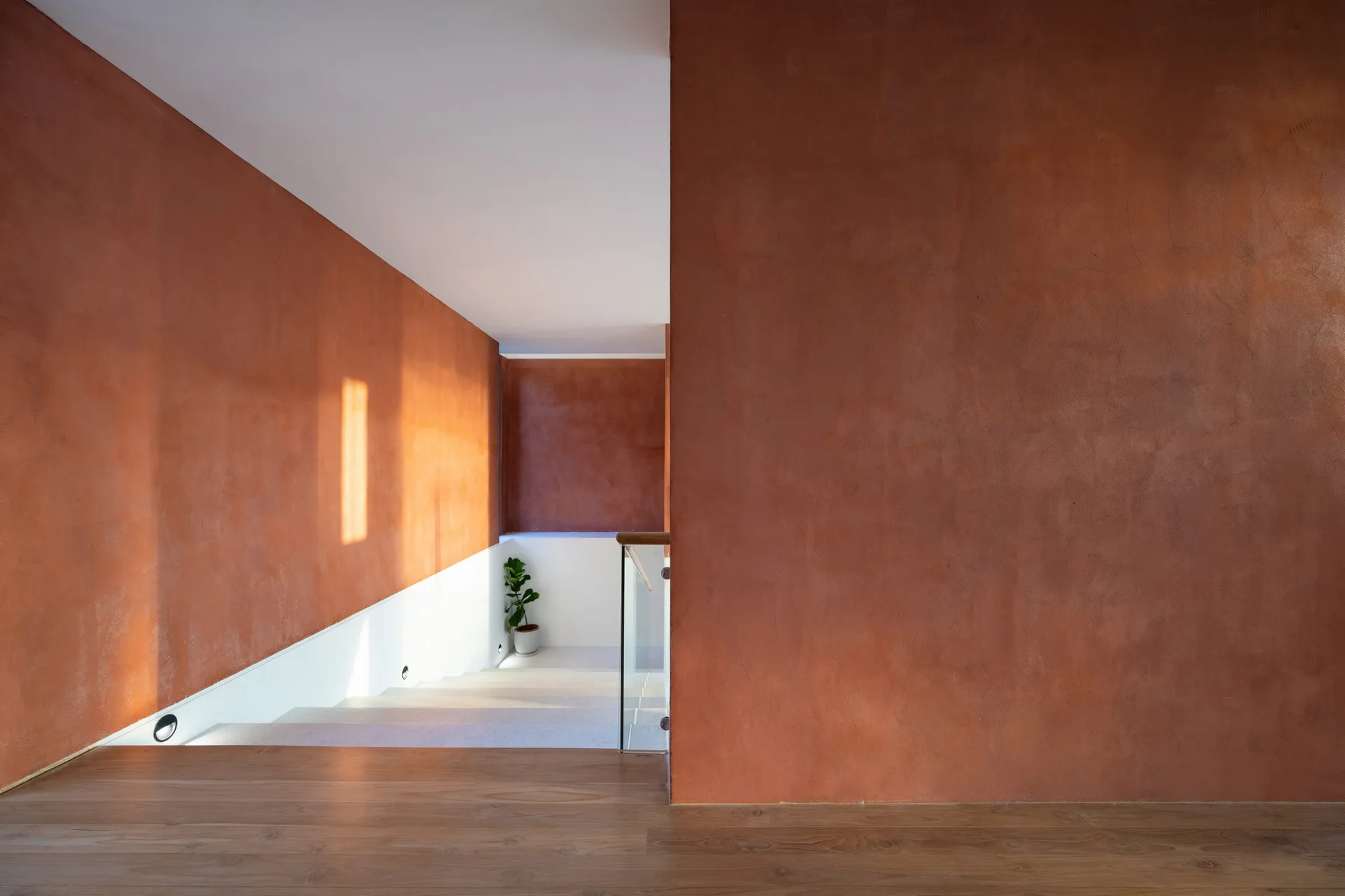 A minimalist residential staircase features textured terracotta plaster walls, natural wood flooring, a clear glass railing, and subtle floor lighting.