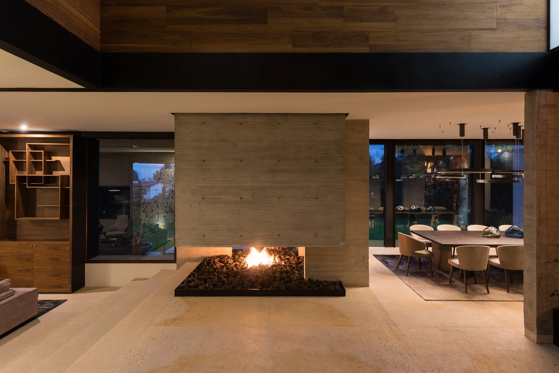 A modern living space featuring a central concrete fireplace partition, wood ceiling paneling, and an adjacent dining area.