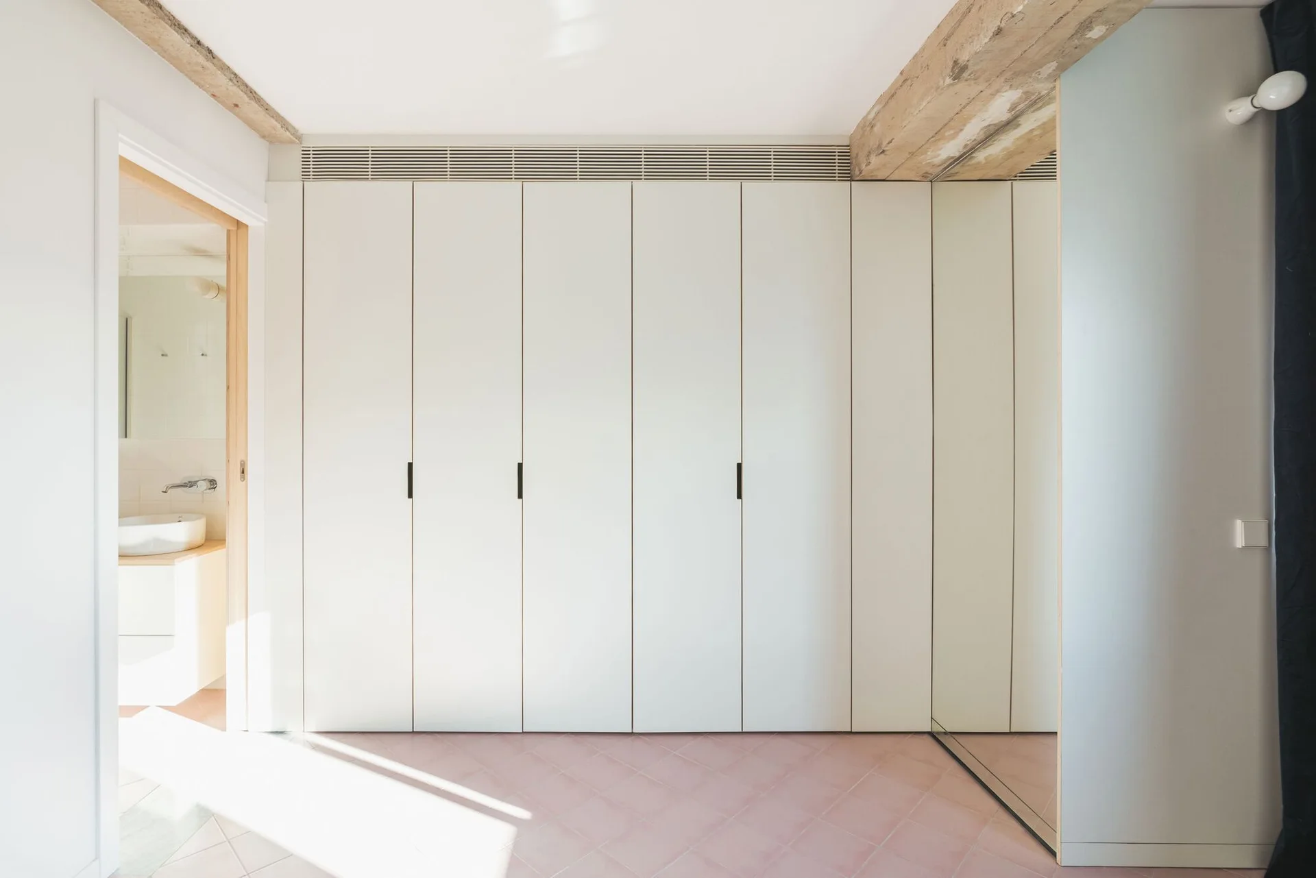 A minimalist off-white built-in closet stands next to an open doorway showing a bathroom vanity, set against pink tiled floors and an exposed wooden beam.
