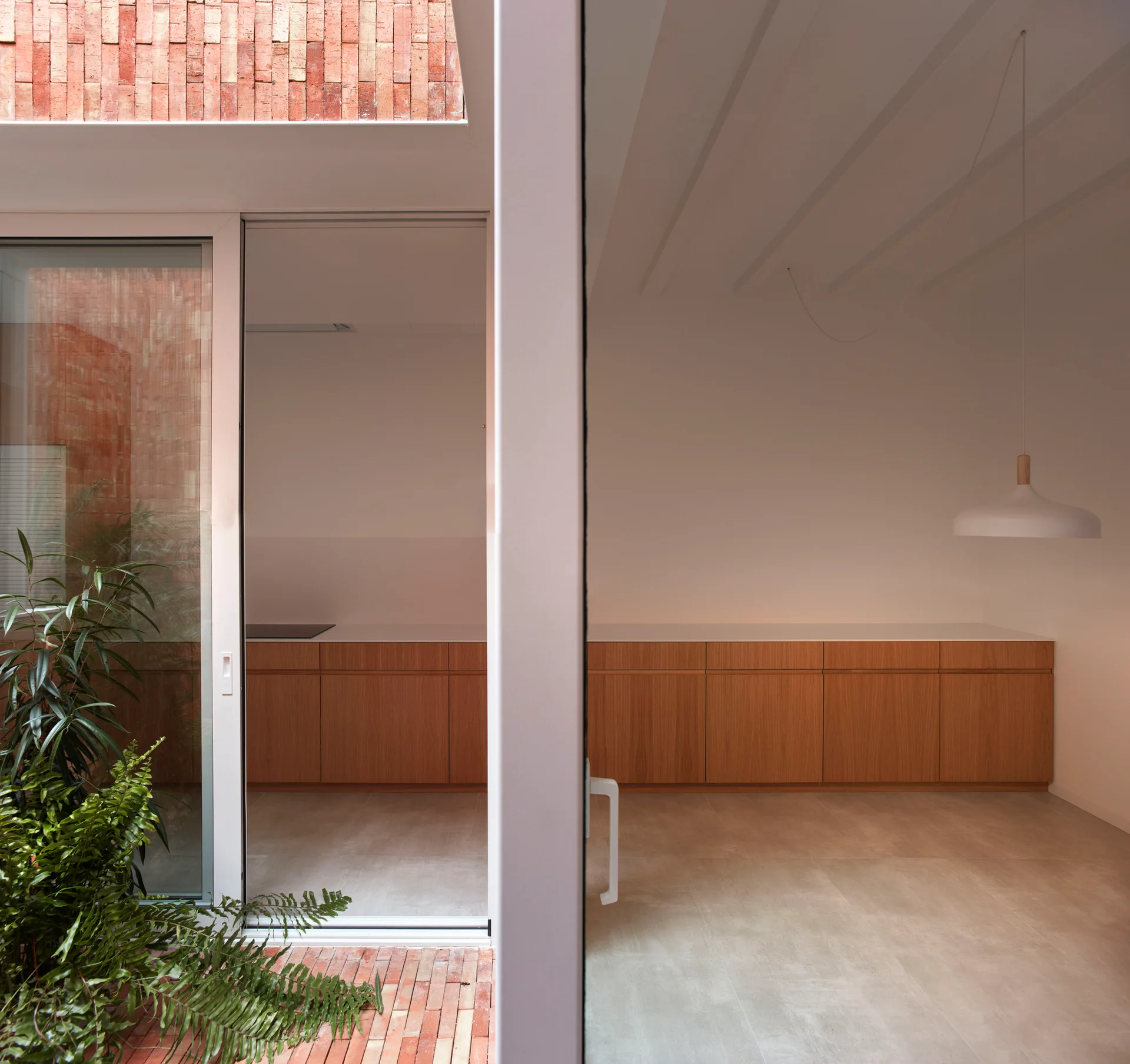 A minimalist kitchen features wood cabinets and a sliding glass door opening to an outdoor brick courtyard with green plants.