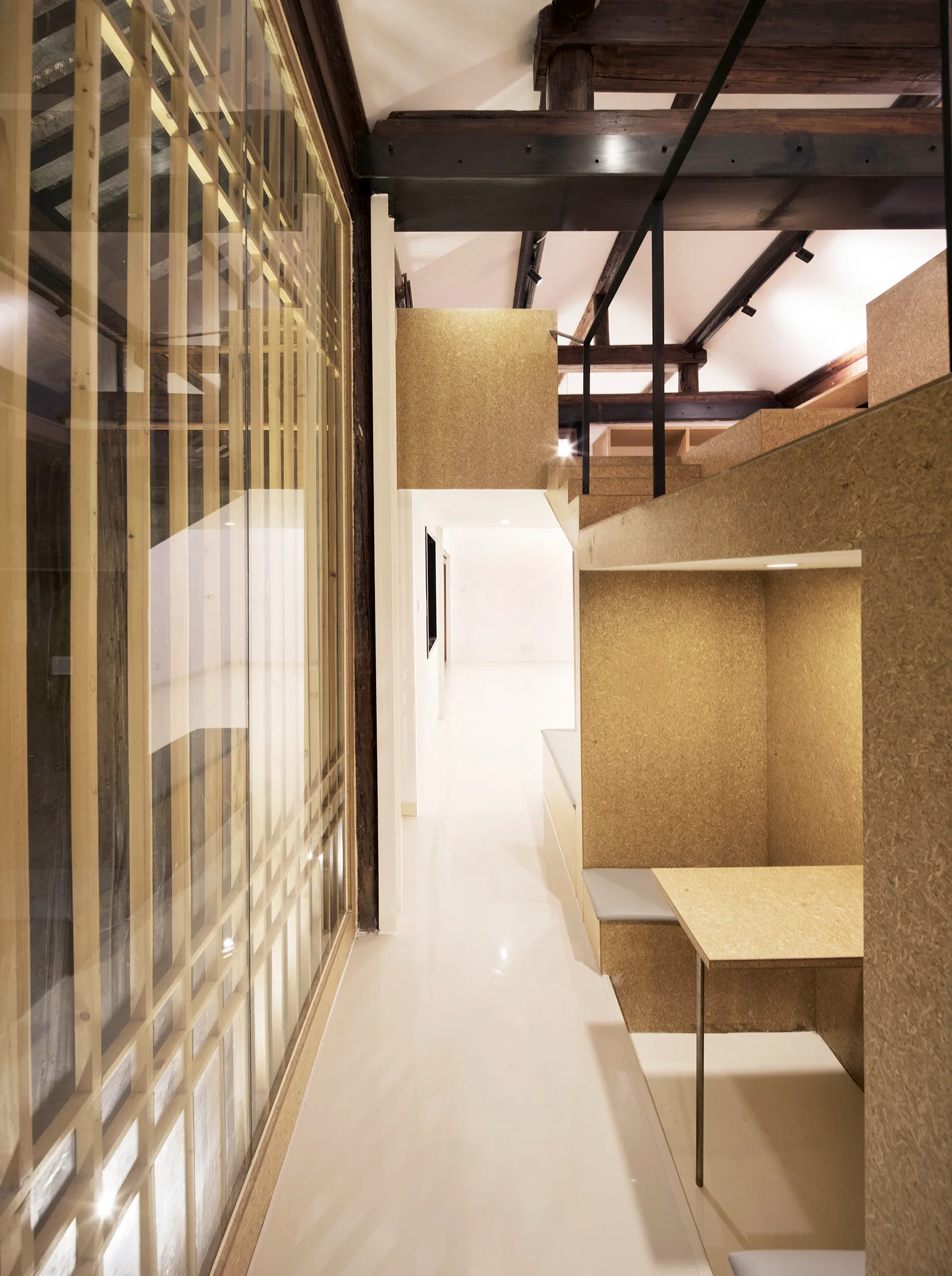 A modern hallway features a glossy white floor, exposed dark timber ceiling beams, a glass partition with a wooden grid, and a built-in OSB wood desk nook.