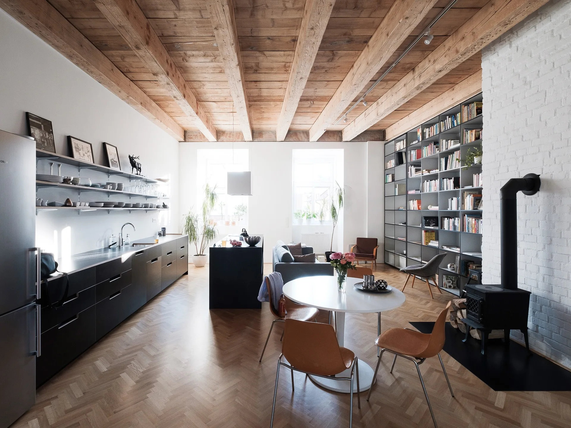 An open-concept loft space features a black kitchen, a white brick chimney with a wood stove, and a high ceiling with exposed wood beams.