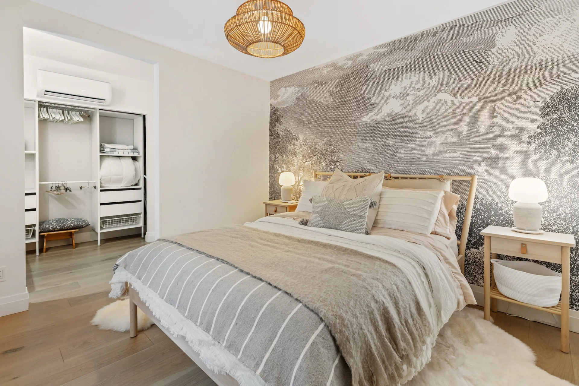 A bedroom featuring a bed with layered textiles set against a gray landscape wall mural, flanked by wooden nightstands with white table lamps.