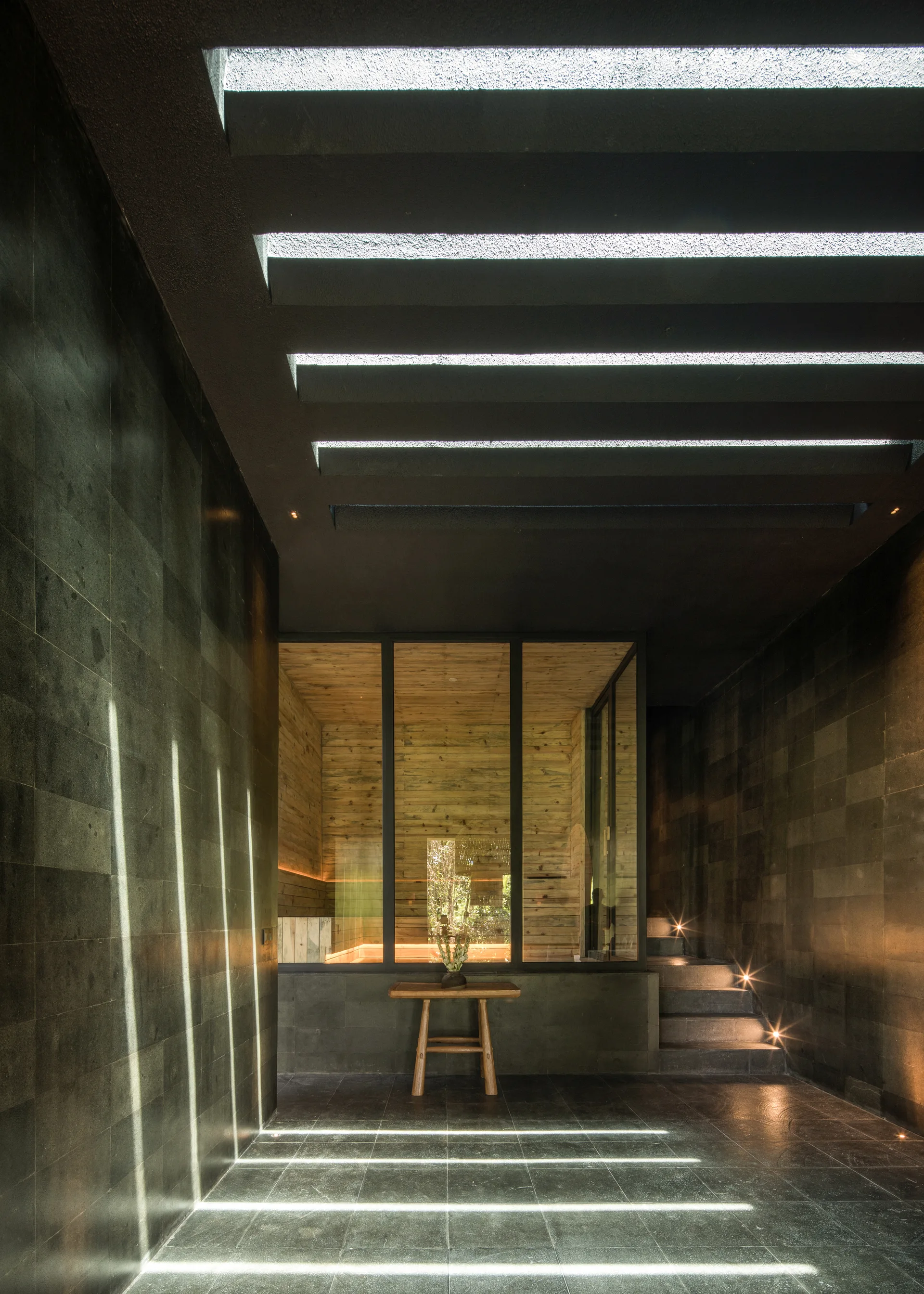 A dark stone corridor features a slotted ceiling casting parallel light strips across the floor, with a wood-lined room visible through a glass partition.