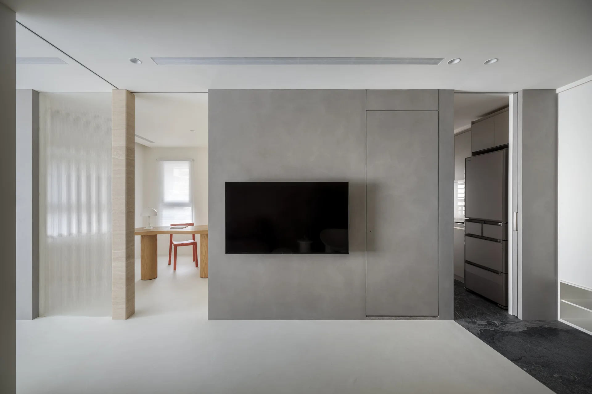 A minimalist living space features a central matte grey media wall with a mounted television and a concealed door, flanked by kitchen and dining areas.