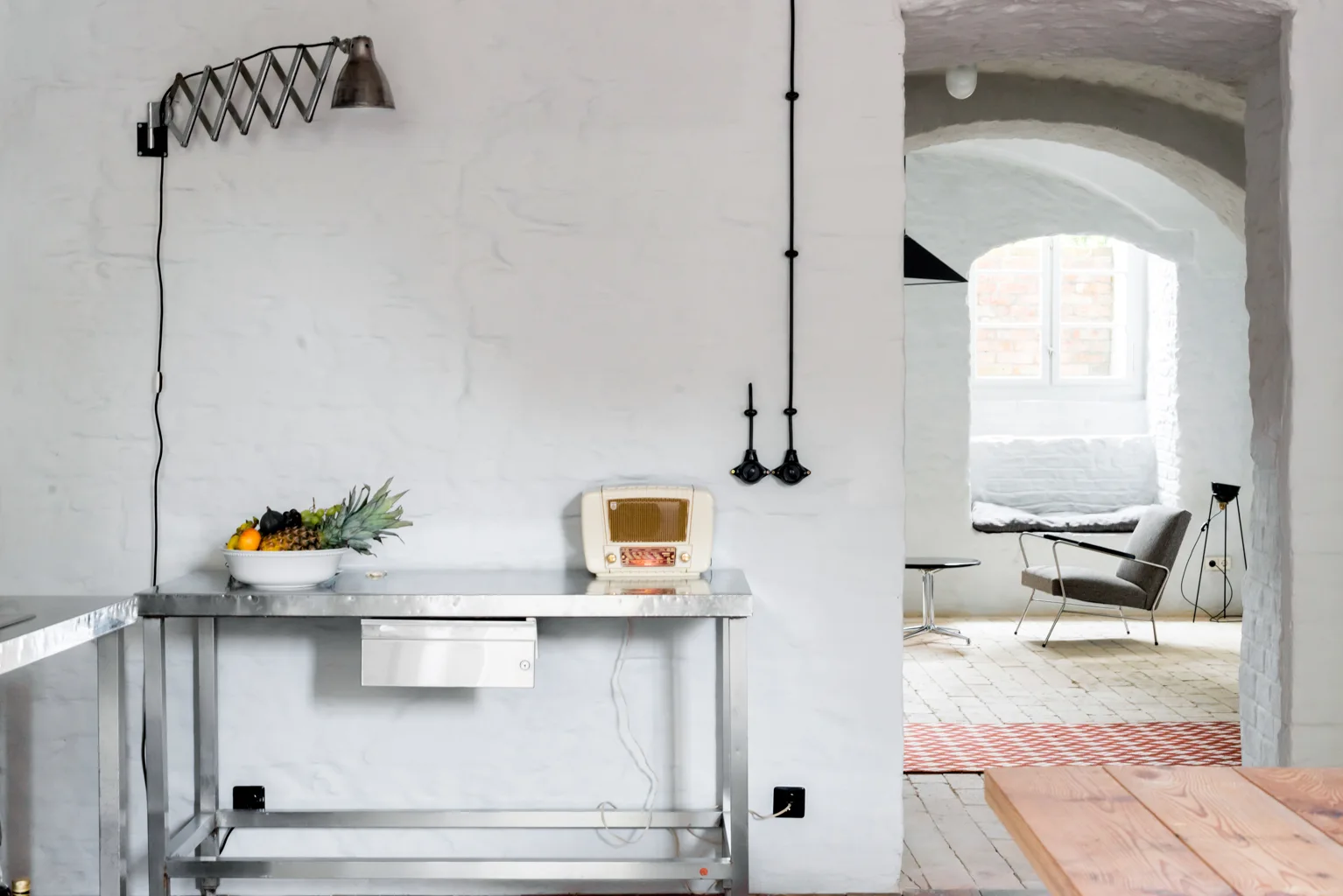 A stainless steel work table with a vintage radio stands against a whitewashed brick wall beneath an accordion lamp, looking into an adjacent room.