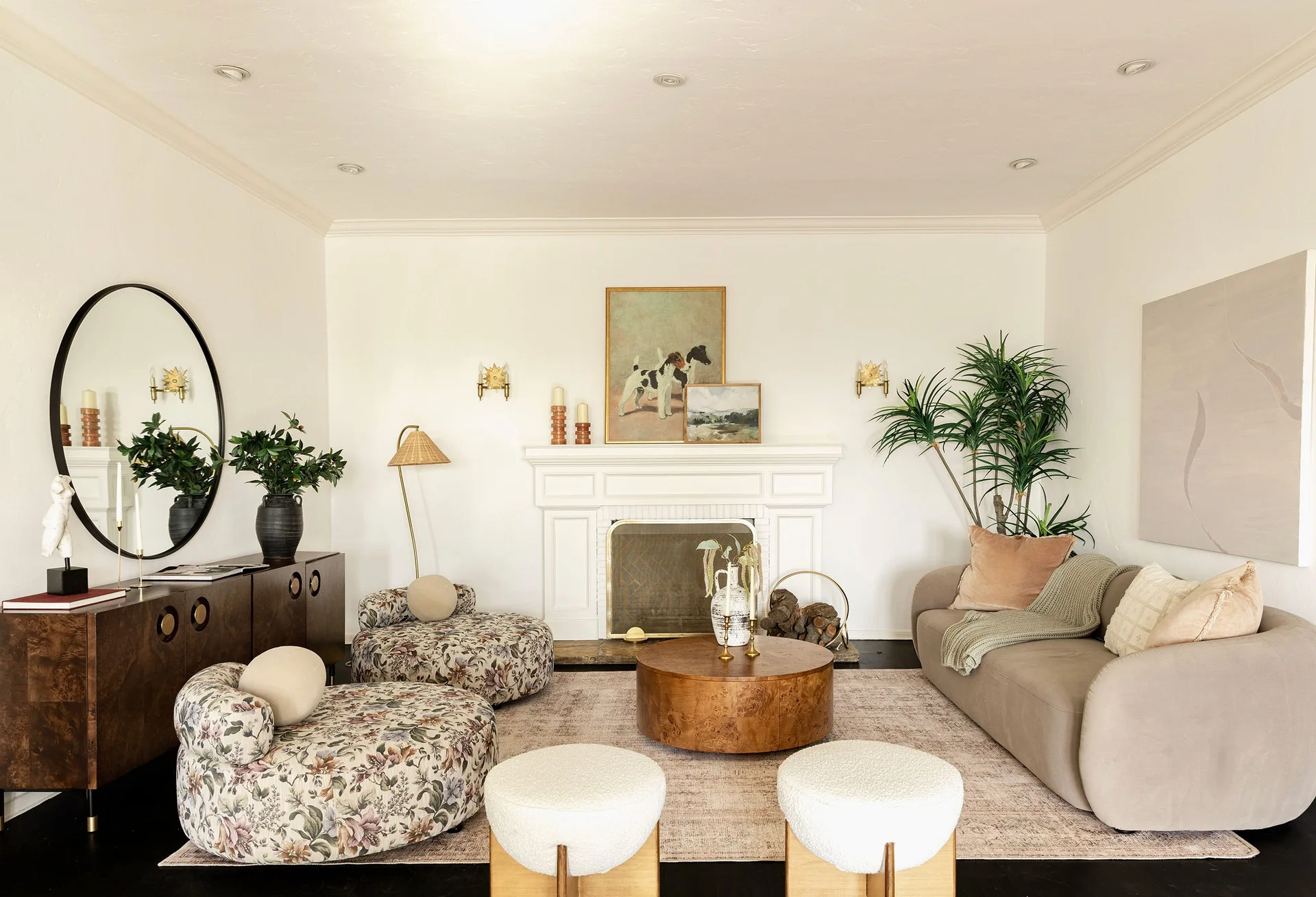 A bright living room featuring a white fireplace mantel, a curved beige sofa, two floral lounge chairs, and a round wooden coffee table.
