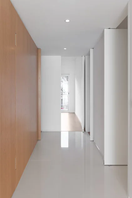 A modern minimalist hallway featuring a floor-to-ceiling wood-paneled wall on one side, clean white walls, and highly reflective glossy tiling.