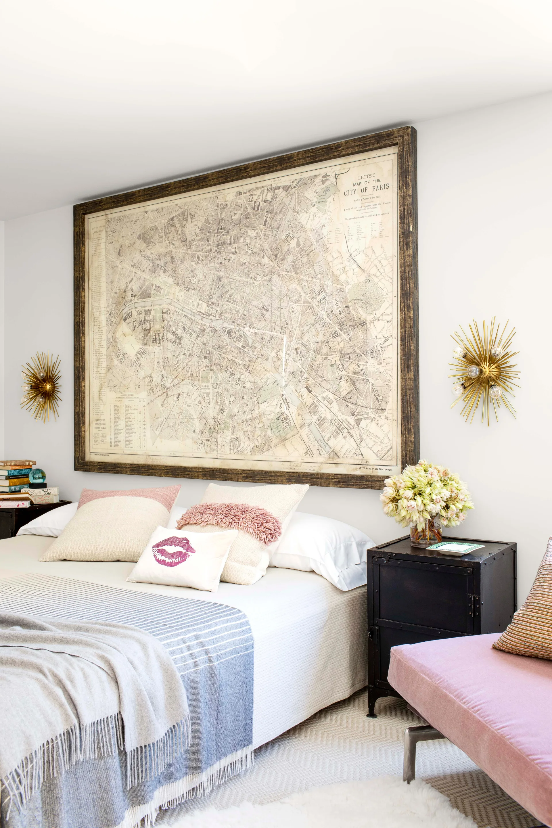 A bright bedroom featuring a large framed vintage map on a white wall above a bed with decorative pillows, flanked by gold starburst sconces.