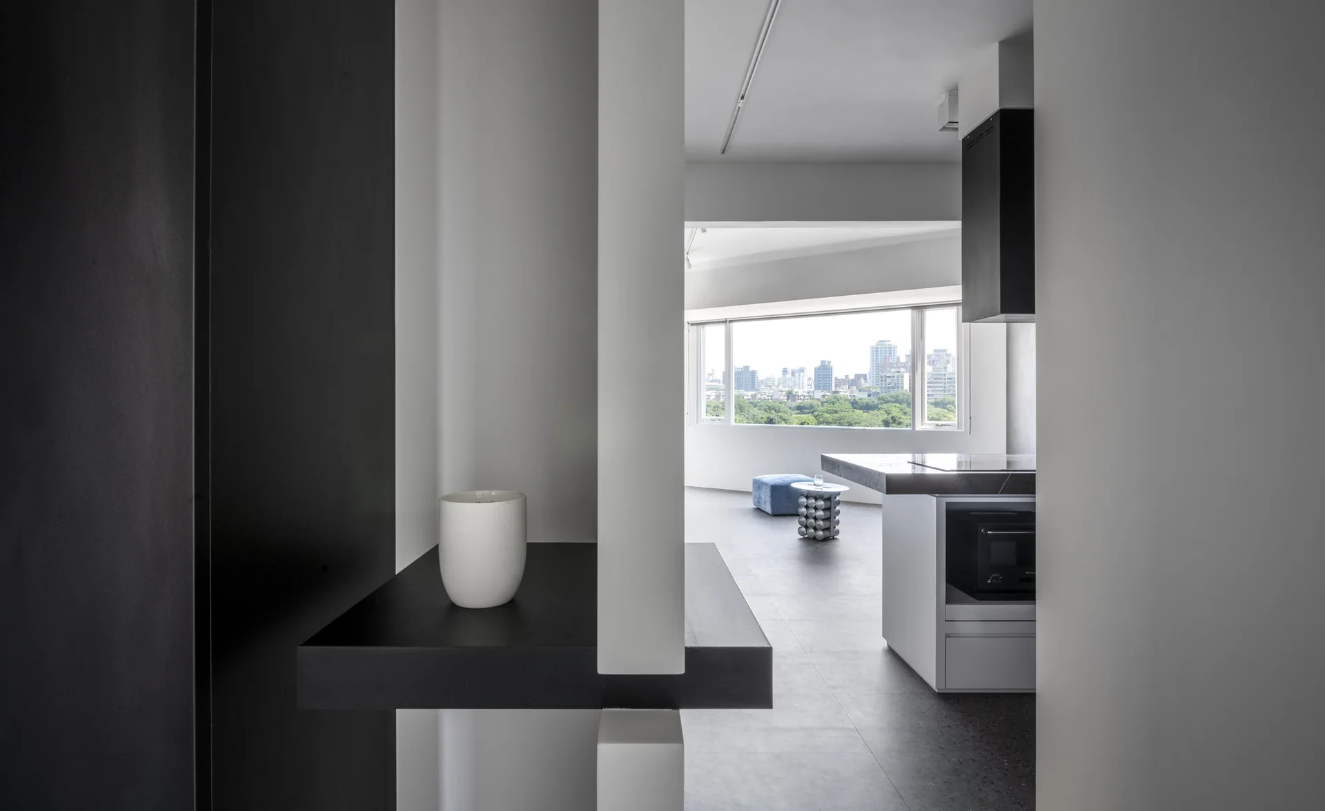 A view through minimalist black and white walls to a modern kitchen island and a bright living area with a large window overlooking a city.