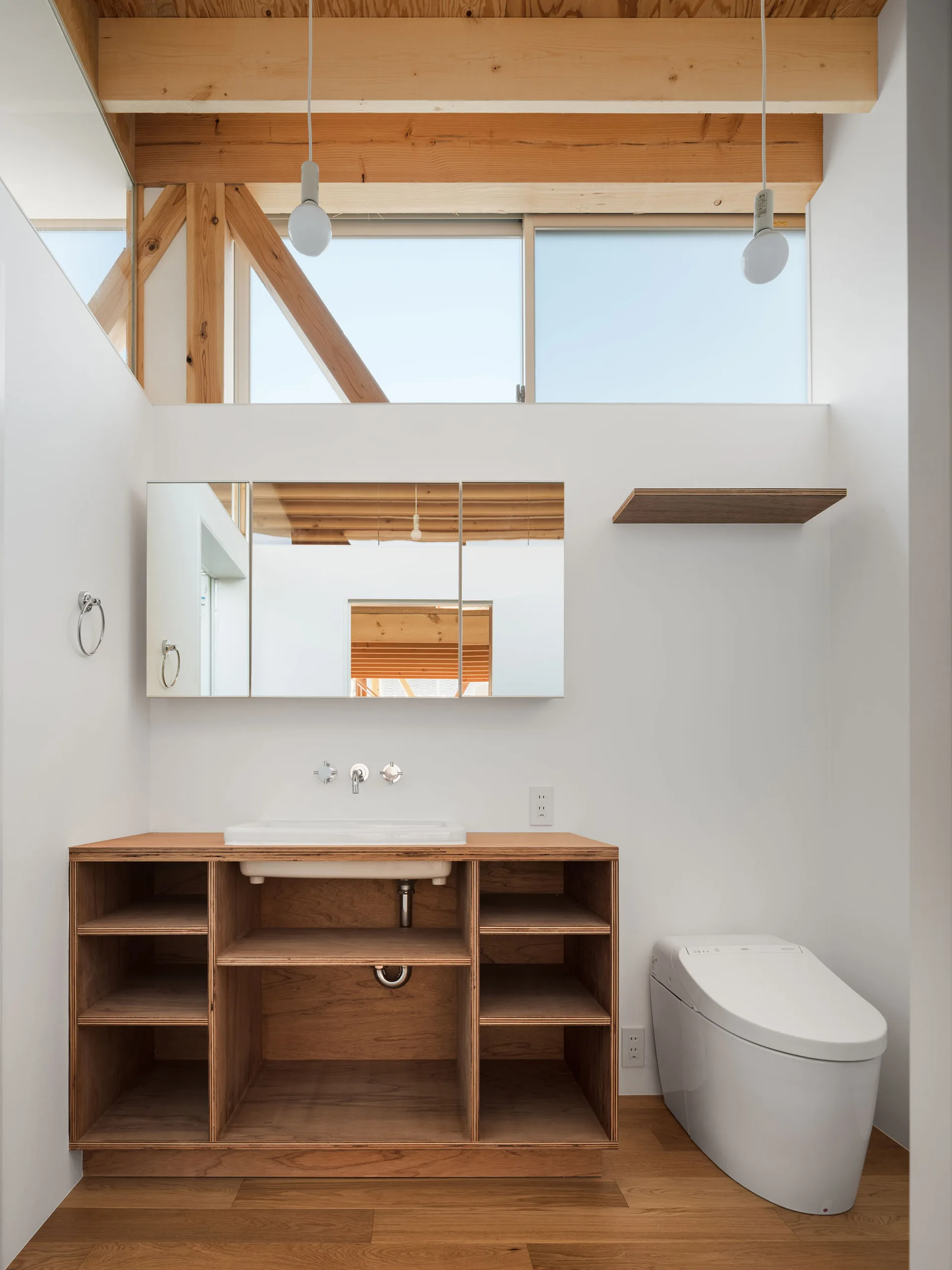 A minimalist bathroom features an open wood vanity with a white sink, a modern toilet, smooth white walls, and exposed timber rafters overhead.