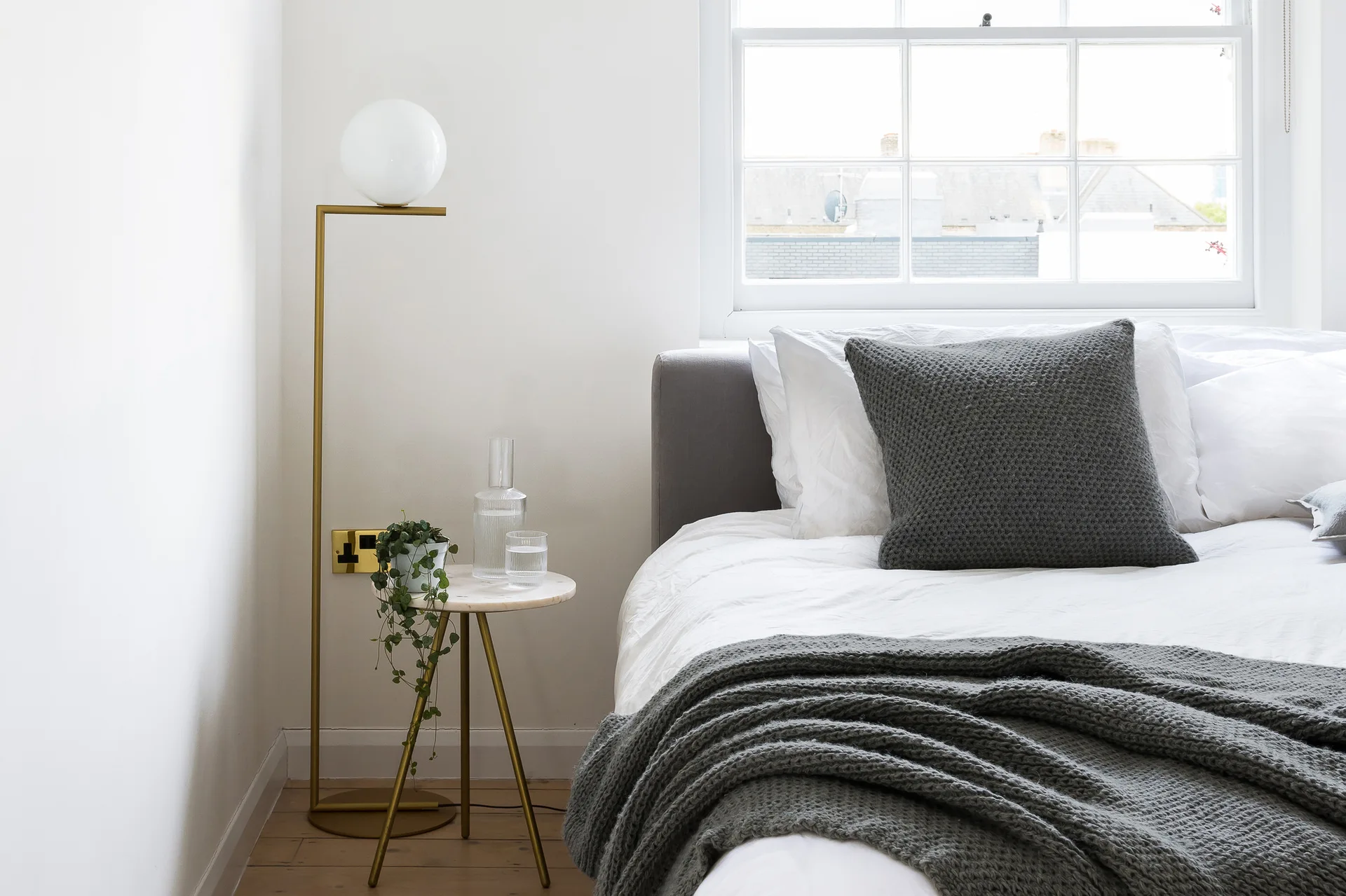 A bright bedroom featuring a bed with grey knit textiles next to a marble-topped side table and a modern brass floor lamp.