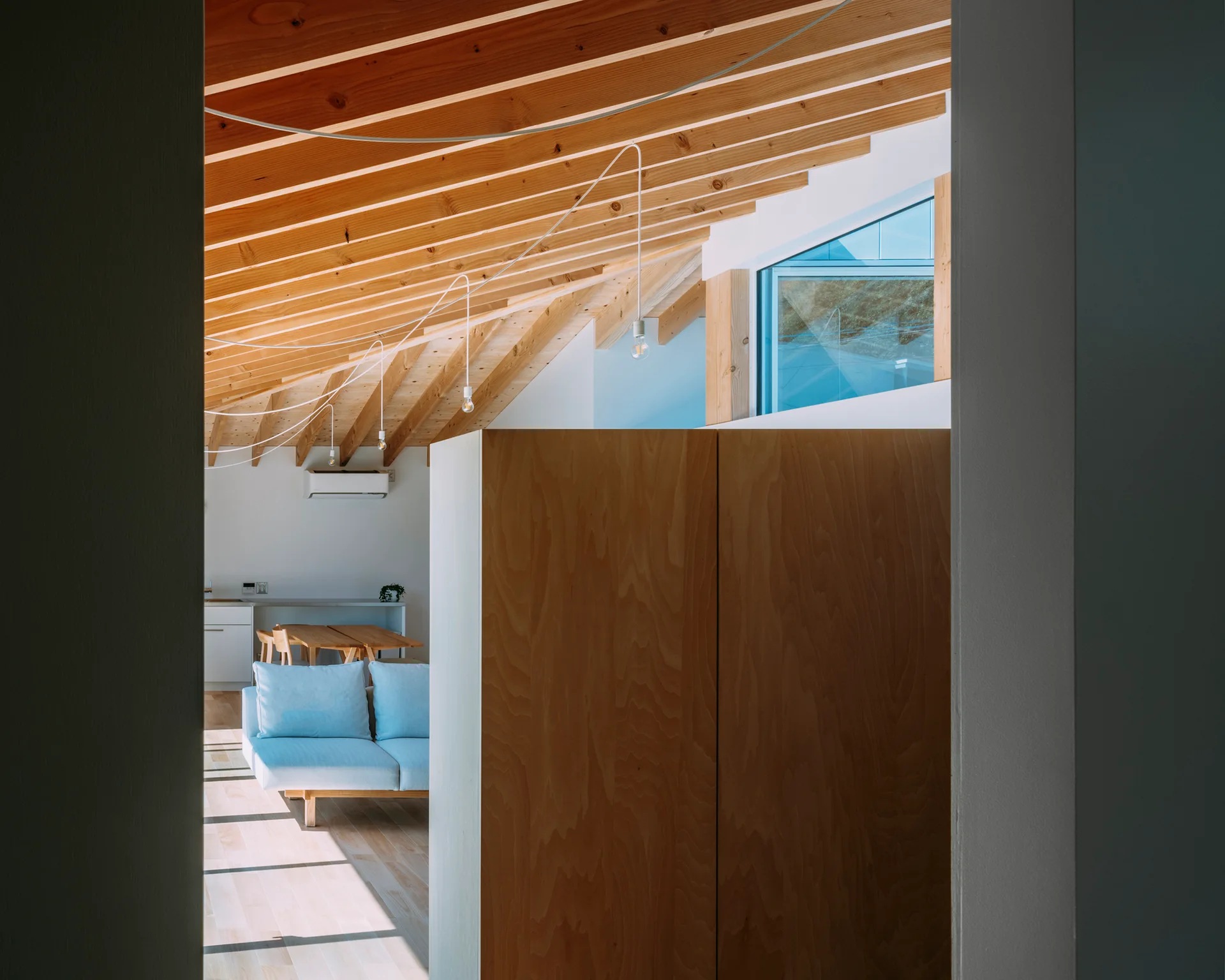 A view through a doorway reveals a bright living room with exposed wood ceiling rafters, a large plywood cabinet, and a light blue sofa.