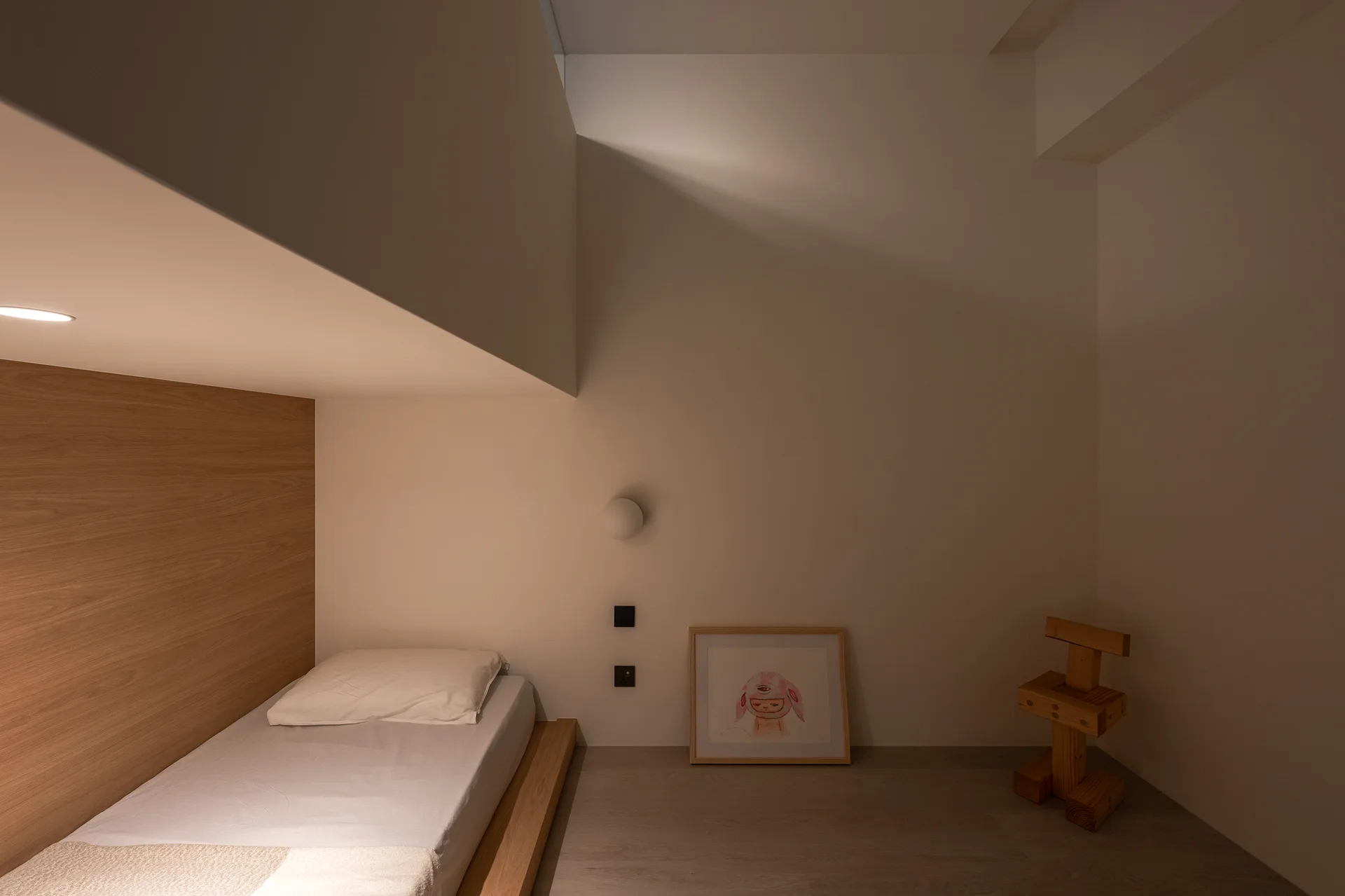 A minimalist bedroom featuring a low wooden platform bed set against a wood paneled wall under a low white overhang.