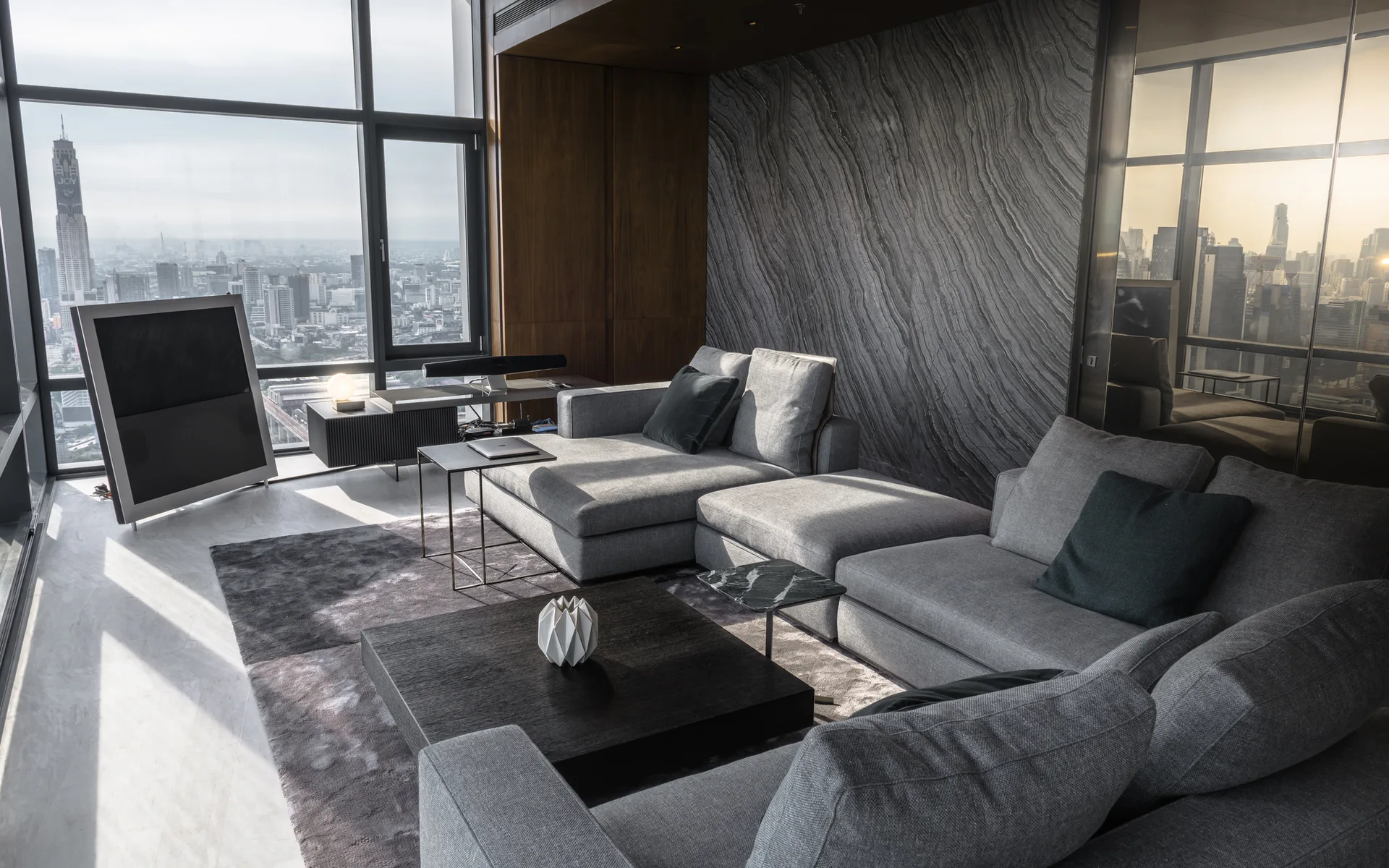 A contemporary living room featuring a large grey sectional sofa, a textured stone feature wall, and windows showing a city skyline.