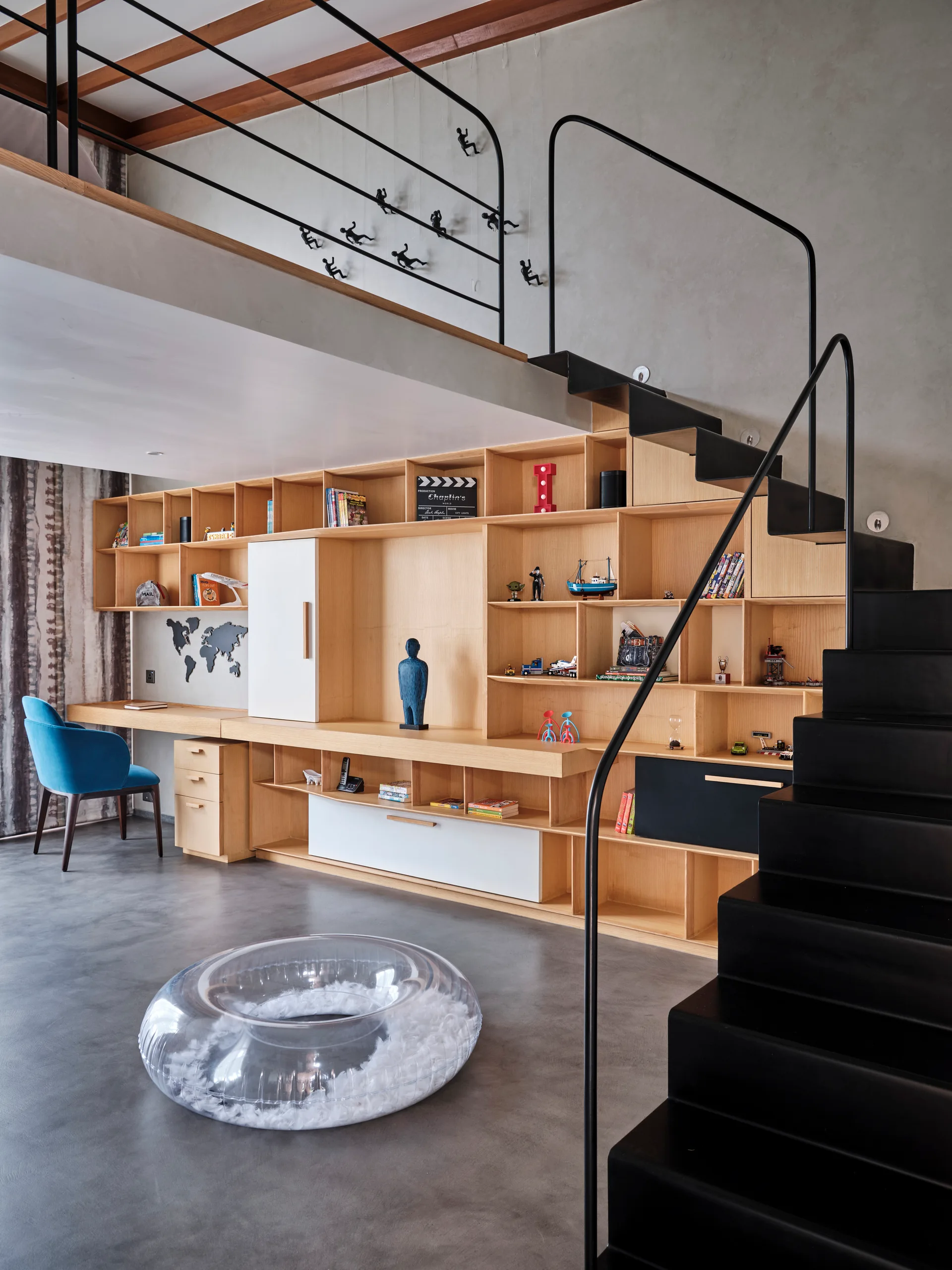 A modern mezzanine home office with a custom light wood shelving unit, built-in desk, concrete floor, and a black steel staircase.