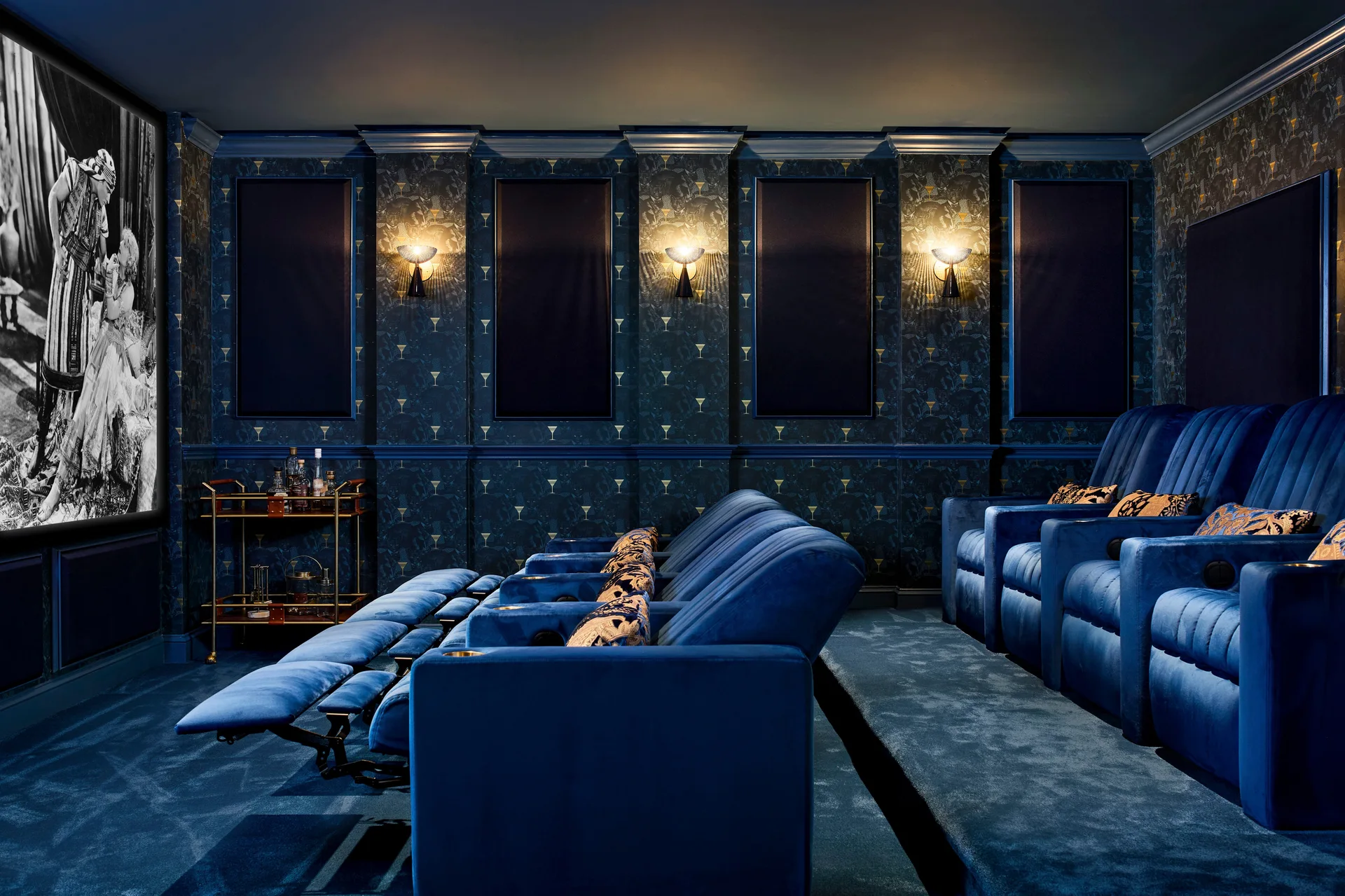 A moody home theater with rows of navy velvet lounge chairs, patterned dark wallpaper, gold sconces, and a large movie screen.