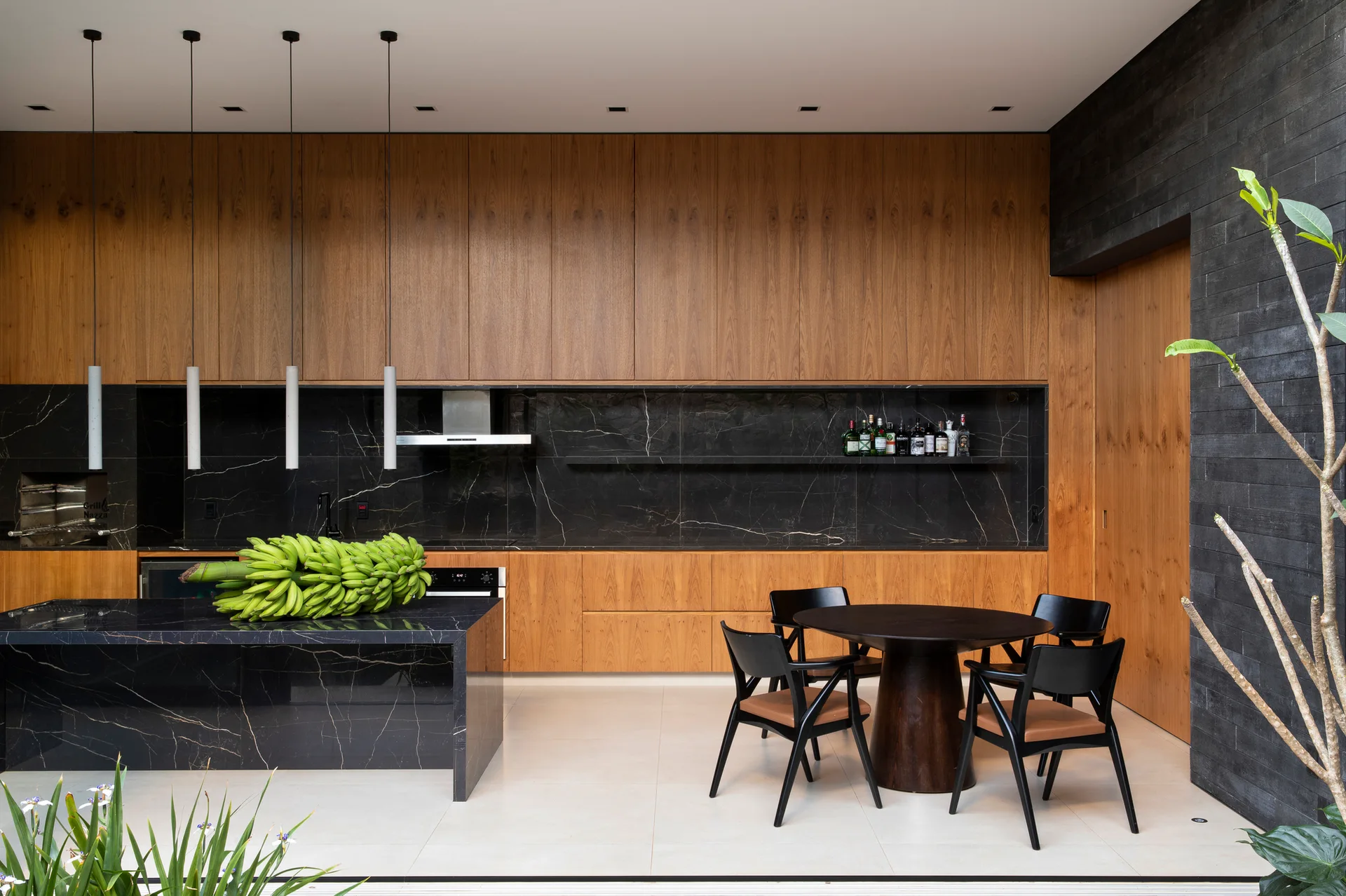 A modern kitchen featuring warm wood cabinetry, a black veined marble waterfall island, and a dark round dining table with four chairs.