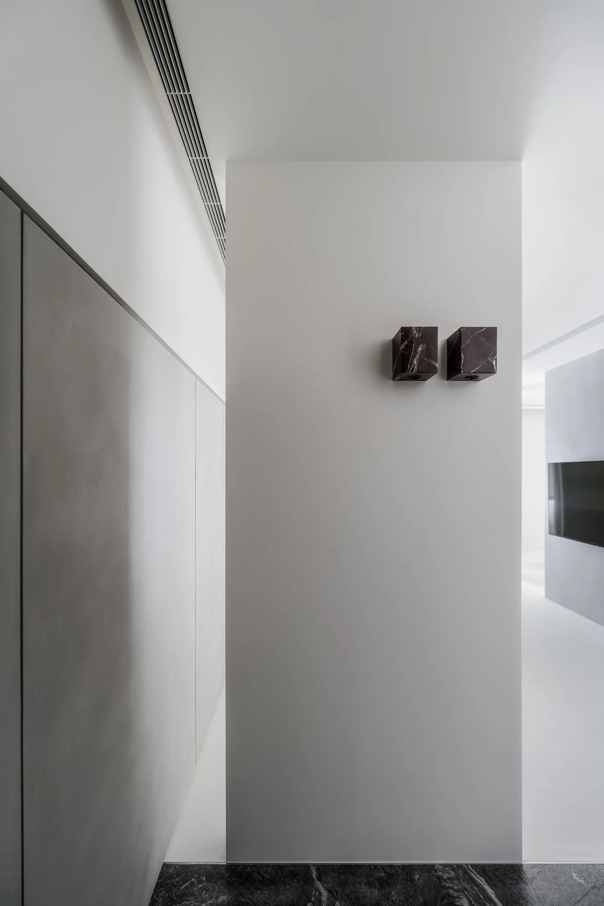 A minimalist hallway features a central white wall section with two dark marble cube sconces, gray wall panels, and a polished black stone floor.