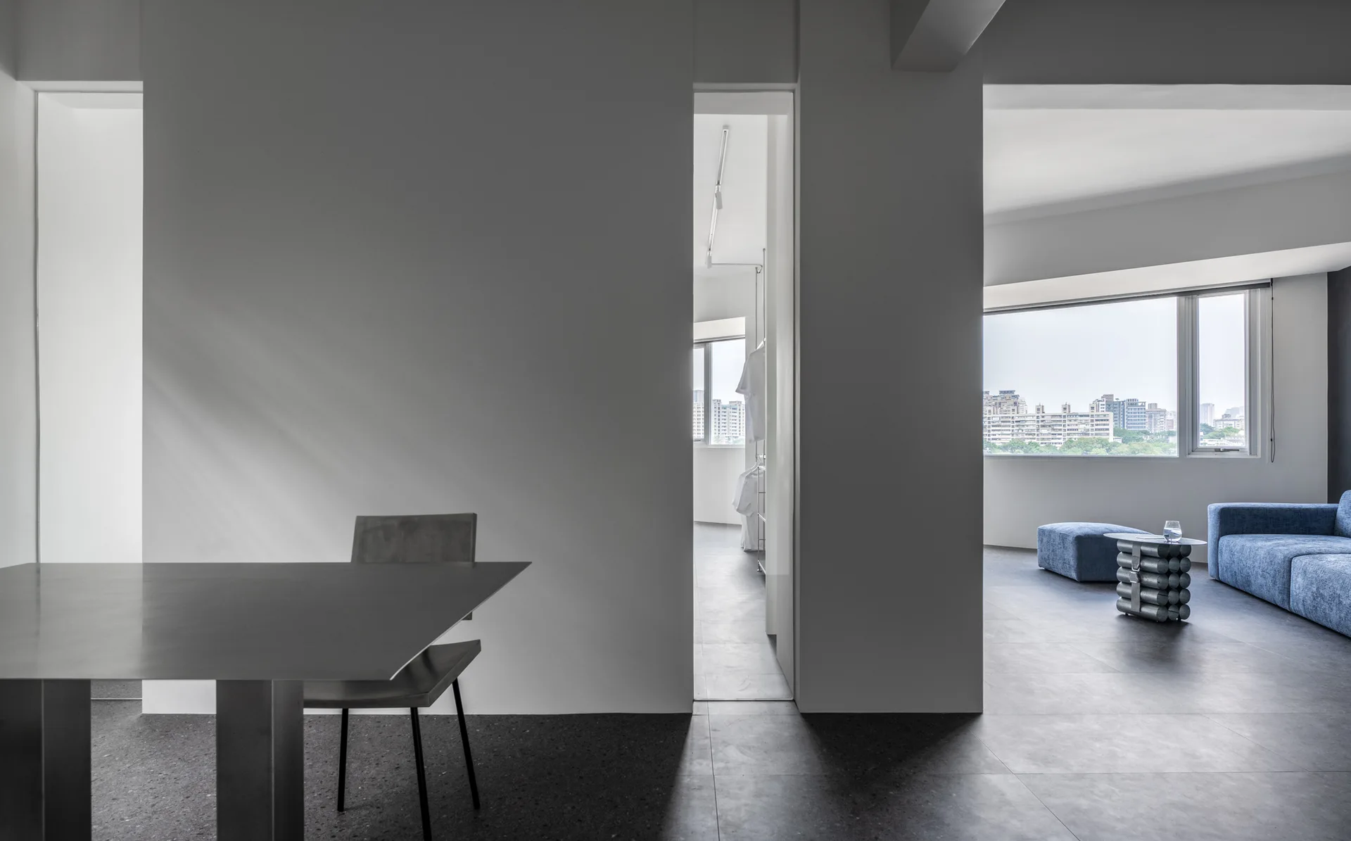 A minimalist grey and white room features a metal table in the foreground, vertical wall openings, and a distant blue sofa near a large window.