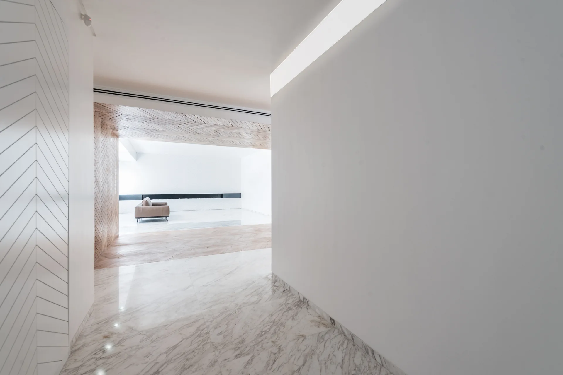 A white minimalist hallway with veined marble tile flooring leads through a herringbone wood archway toward a distant living room.