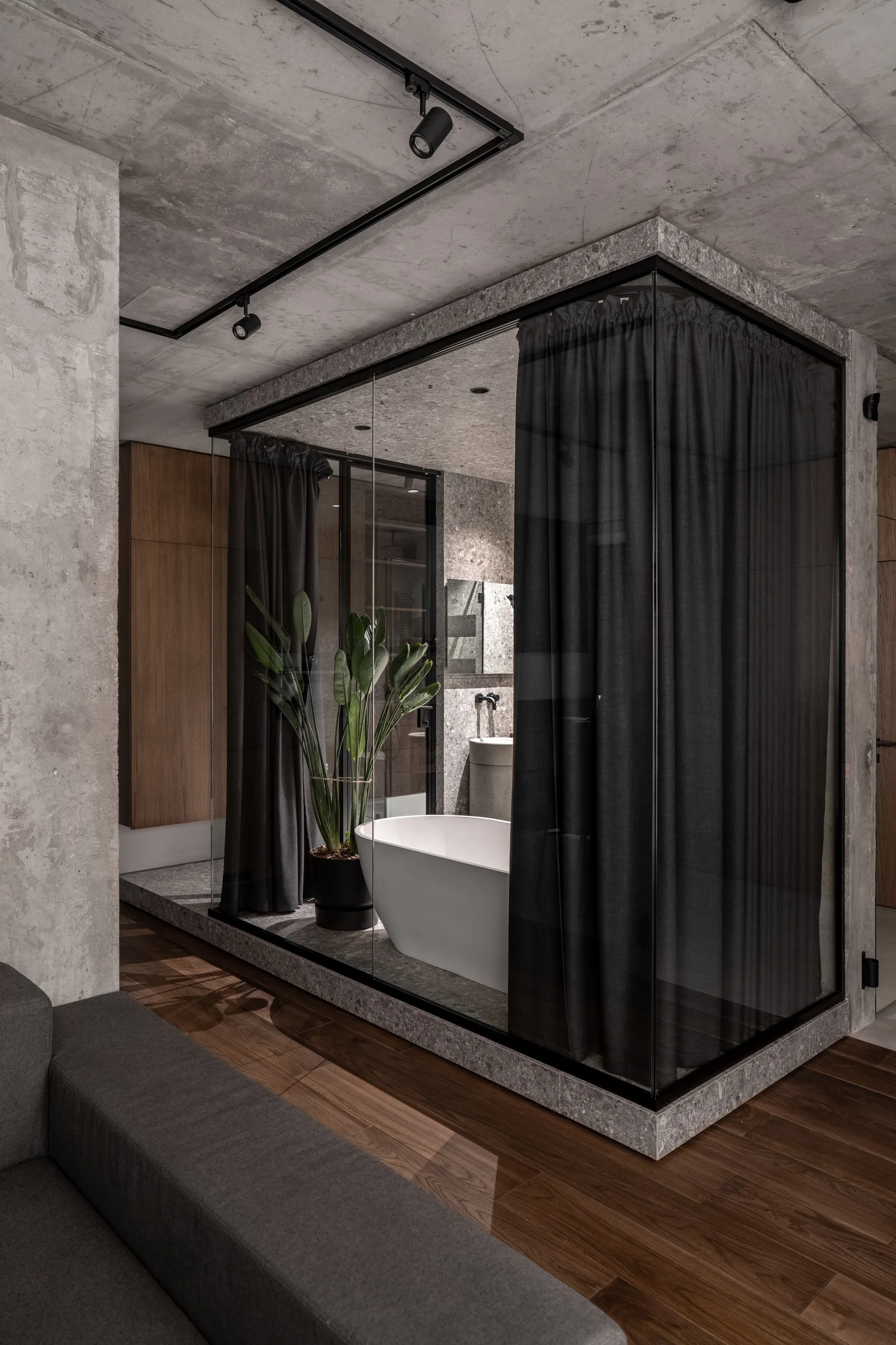 A modern bathroom enclosed in glass walls features a white freestanding bathtub, dark gray curtains, and a raw concrete ceiling with track lighting.