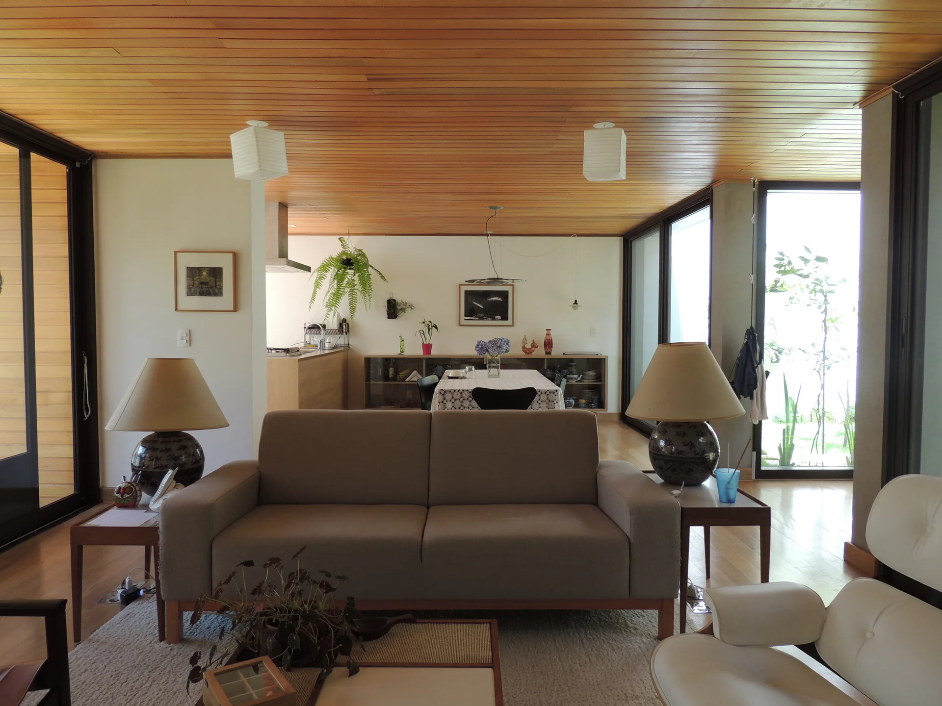 A mid-century modern living room features a beige sofa flanked by table lamps under a wood plank ceiling, with a dining area in the background.