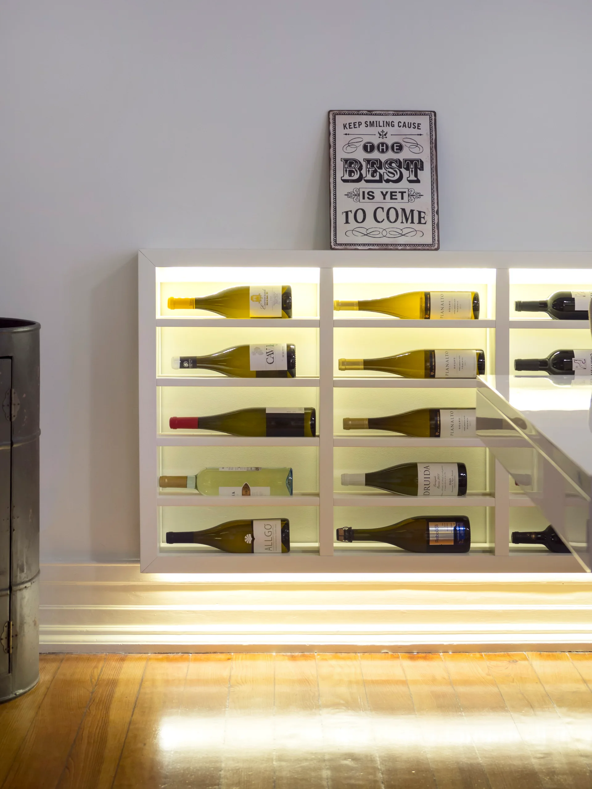 A built-in white wine rack illuminated by warm backlighting holds bottles of wine horizontally above polished hardwood flooring.
